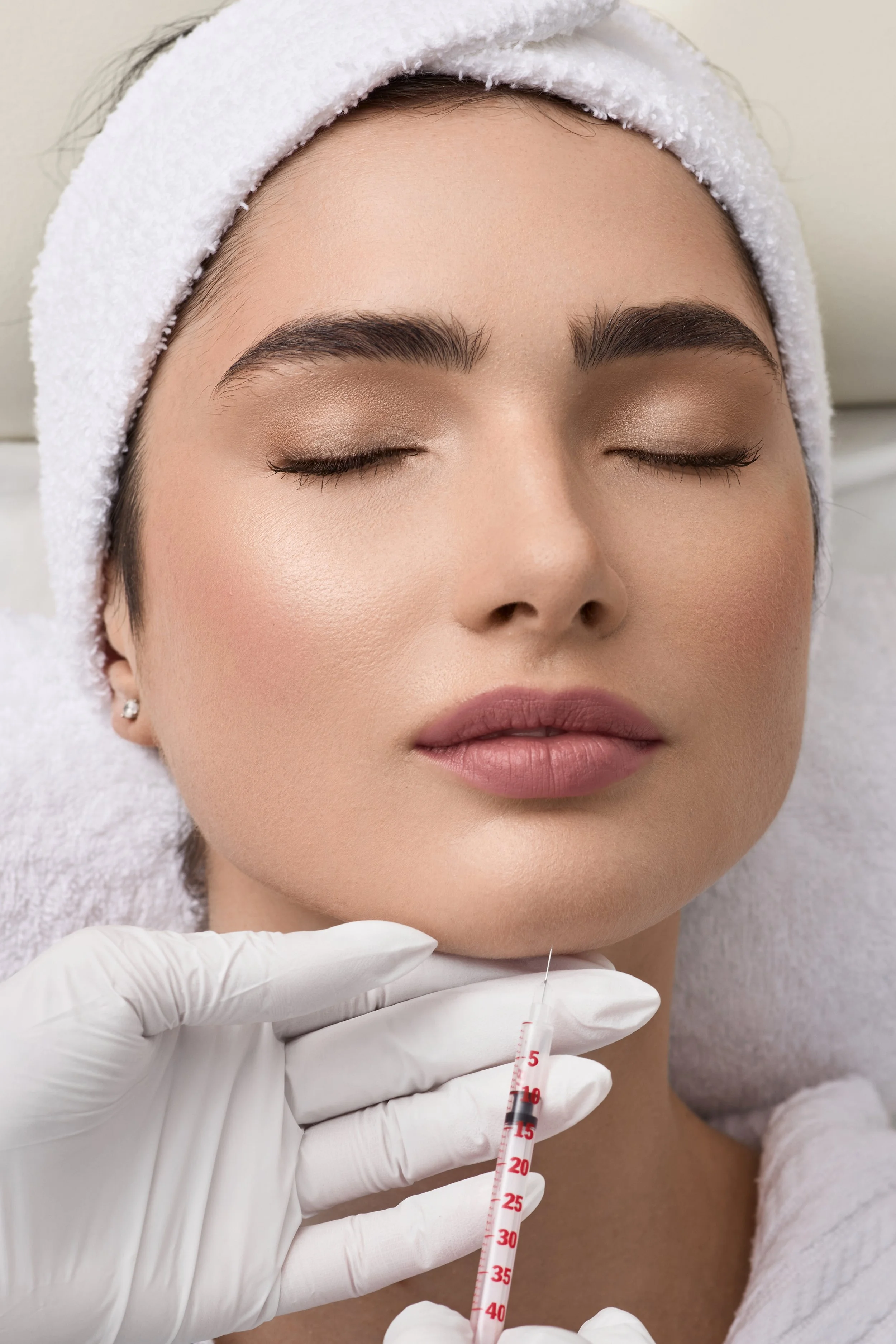 Une femme a les yeux fermés, recevant une injection sous-cutanée dans le menton, dans un contexte médical, avec des gants blancs. Elle porte une serviette blanche autour de la tête.