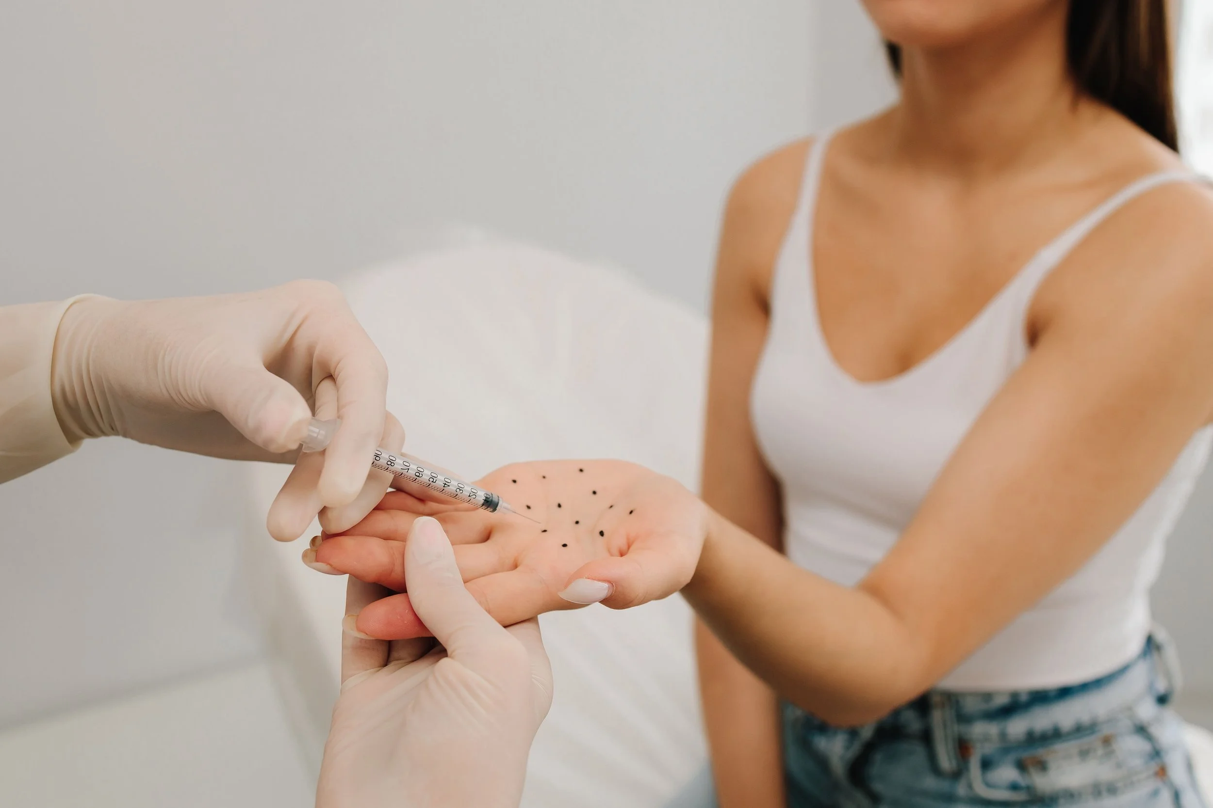 Une femme en débardeur blanc reçoit une injection dans la main d'une personne portant des gants en latex.