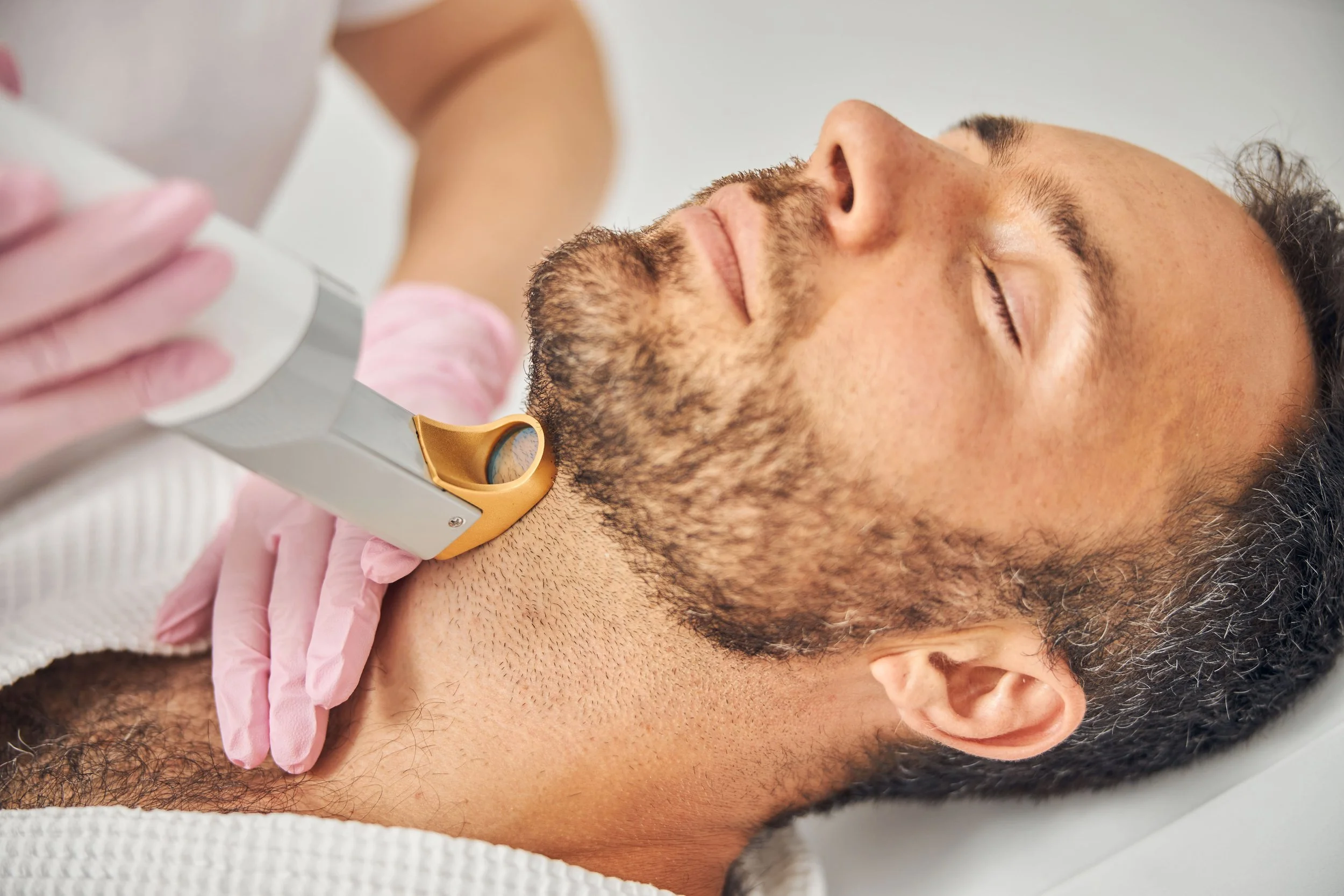Un homme à barbe reçoit un soin du visage avec un appareil de beauté sur son menton, allongé et détendu, porté par une professionnel aux mains gantées roses.