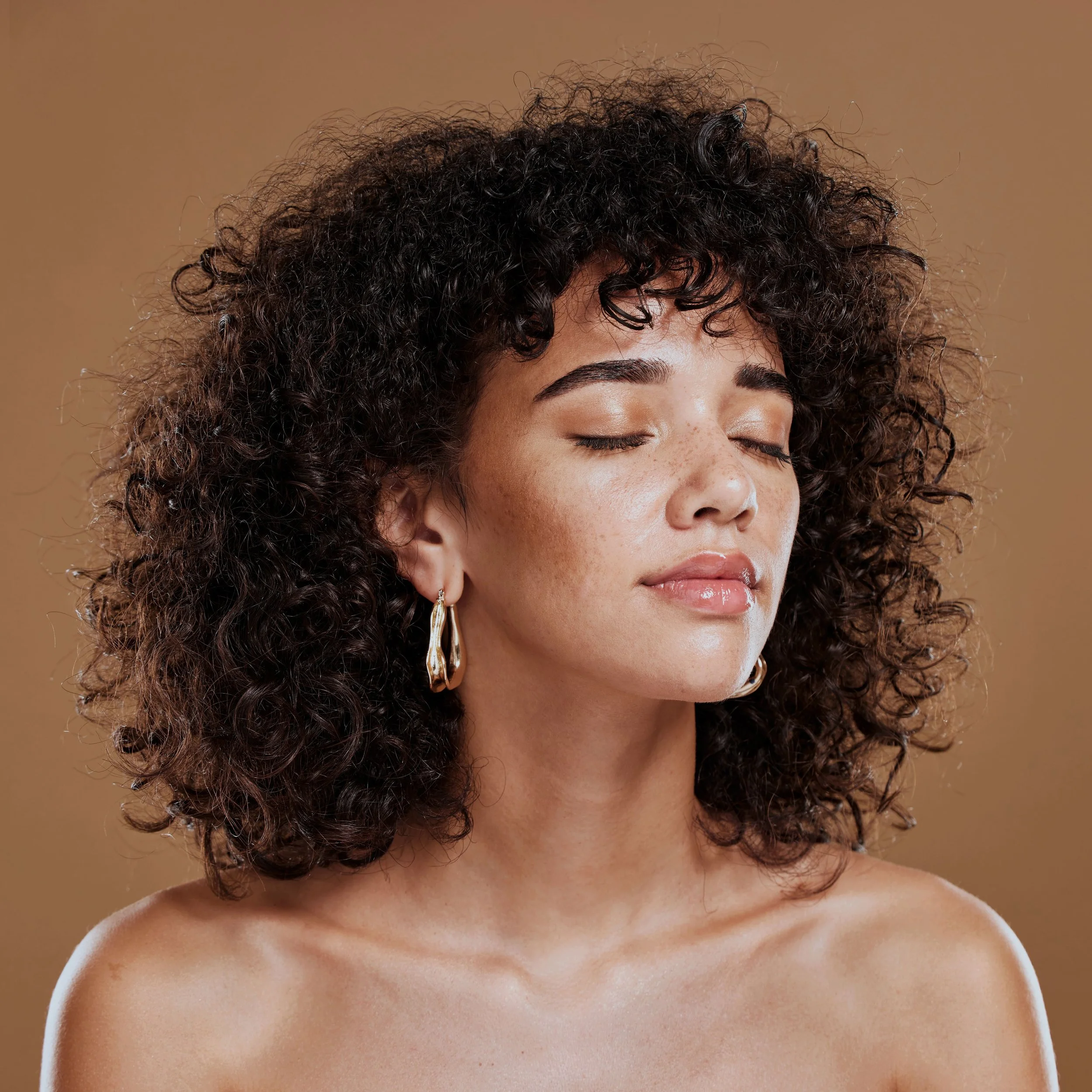 Une femme aux cheveux bouclés, yeux fermés, portant un seul boucle d'oreille, avec une peau humide et un fond beige.