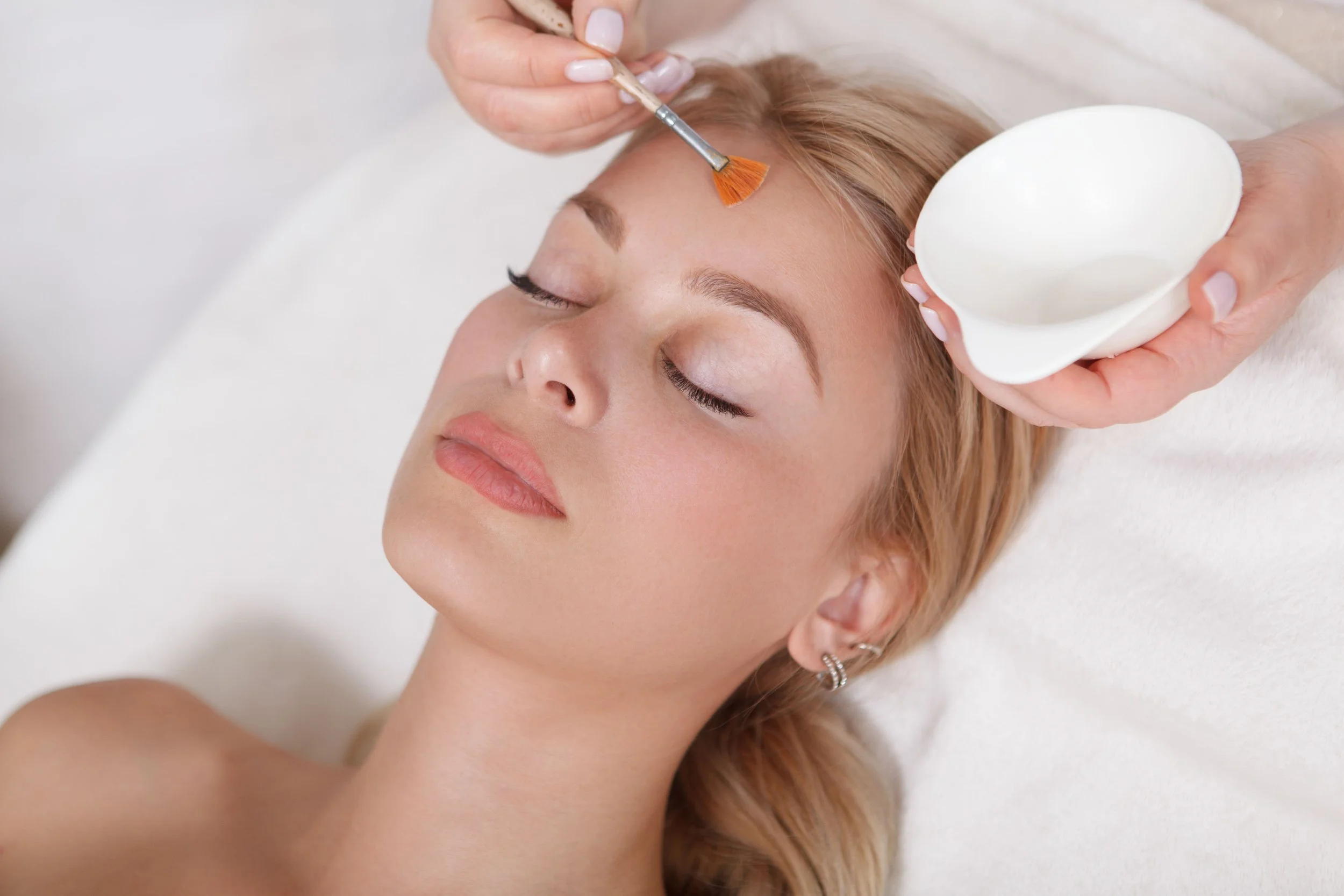 Une femme allongée reçoit un traitement du visage, une esthéticienne applique une crème sur son front.