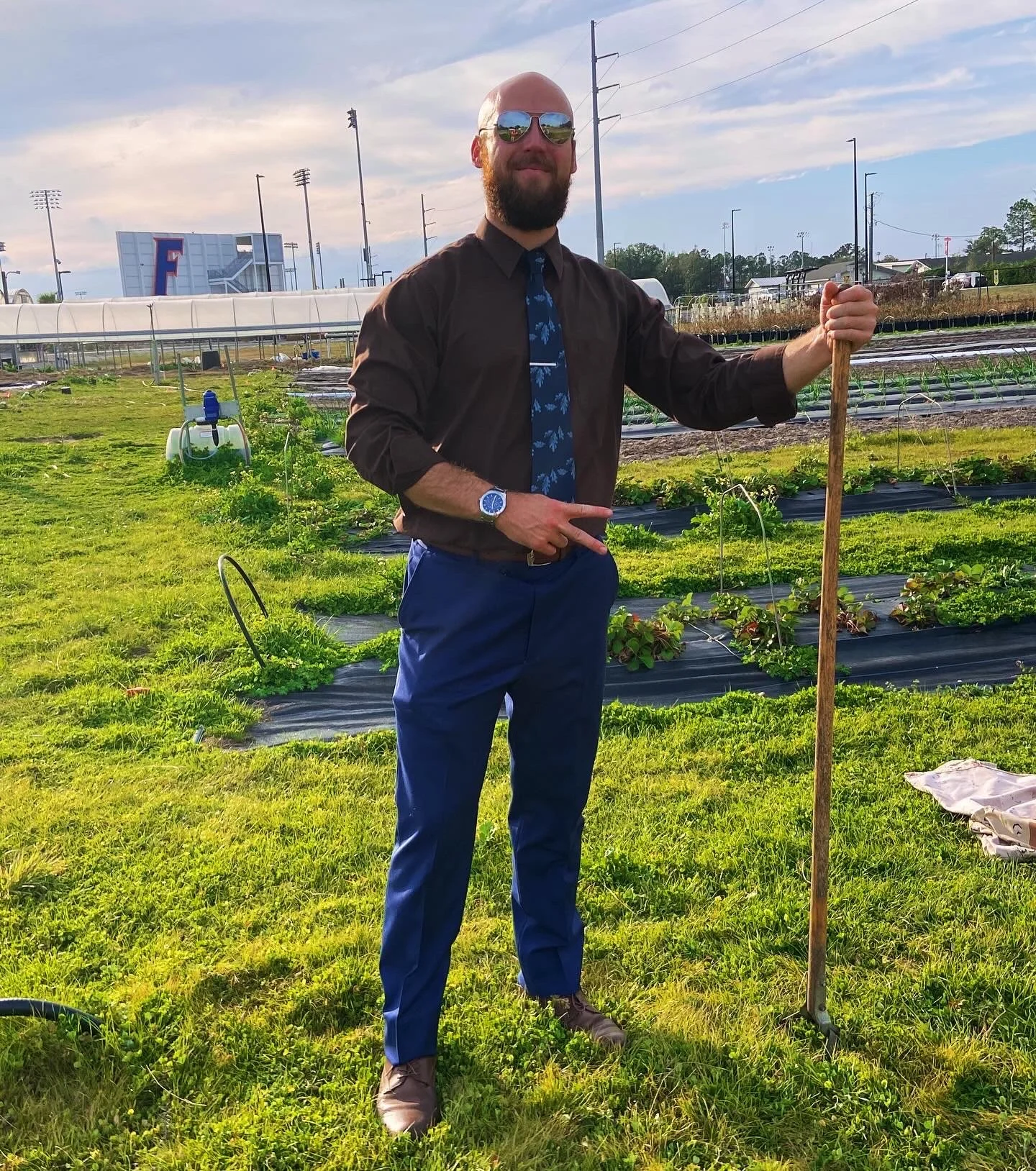 A man with a beard wearing sunglasses, a dark shirt, blue pants, and a patterned tie, stands in a garden holding a gardening tool. The garden has rows of plants covered with mesh, and there are greenhouses and a baseball stadium in the background.