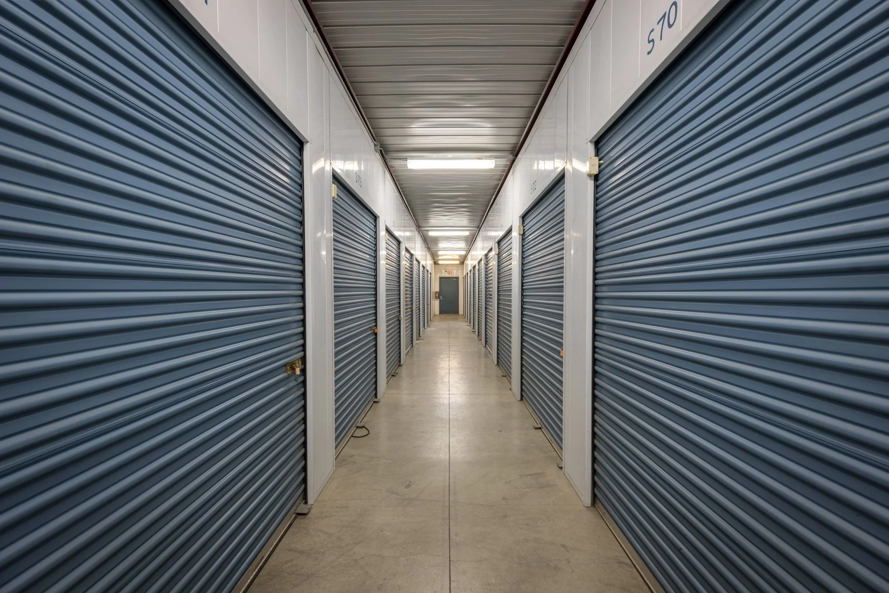 A hallway with rows of storage units with blue roll-up doors on both sides, and fluorescent lighting overhead.