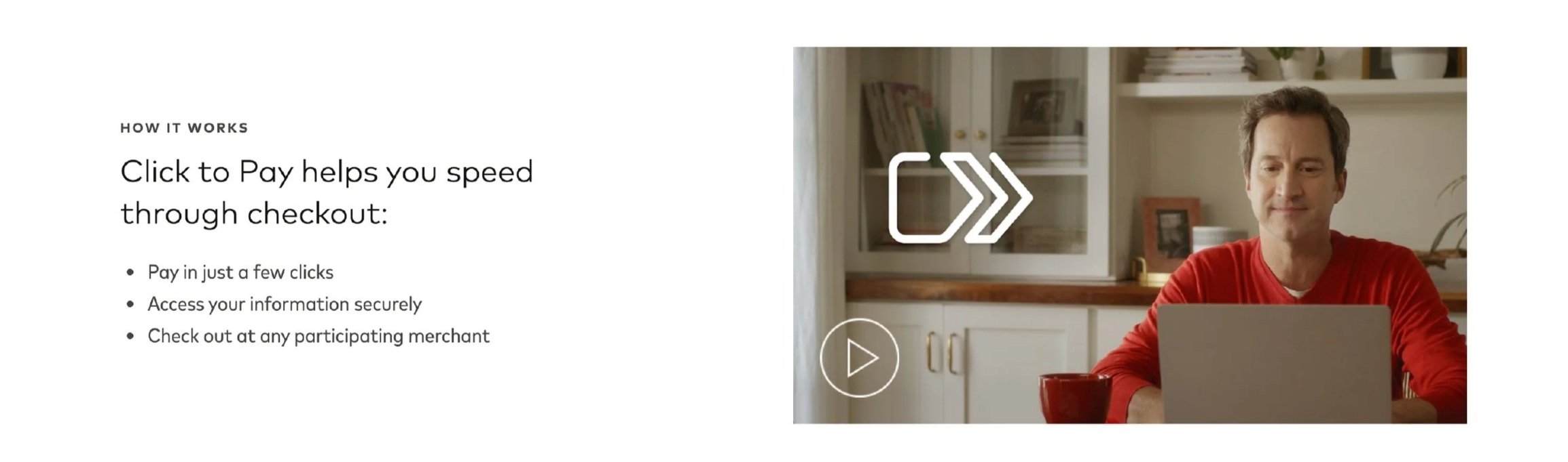 A man in a red shirt using a laptop in a kitchen, with a white shelf and framed photo in the background.