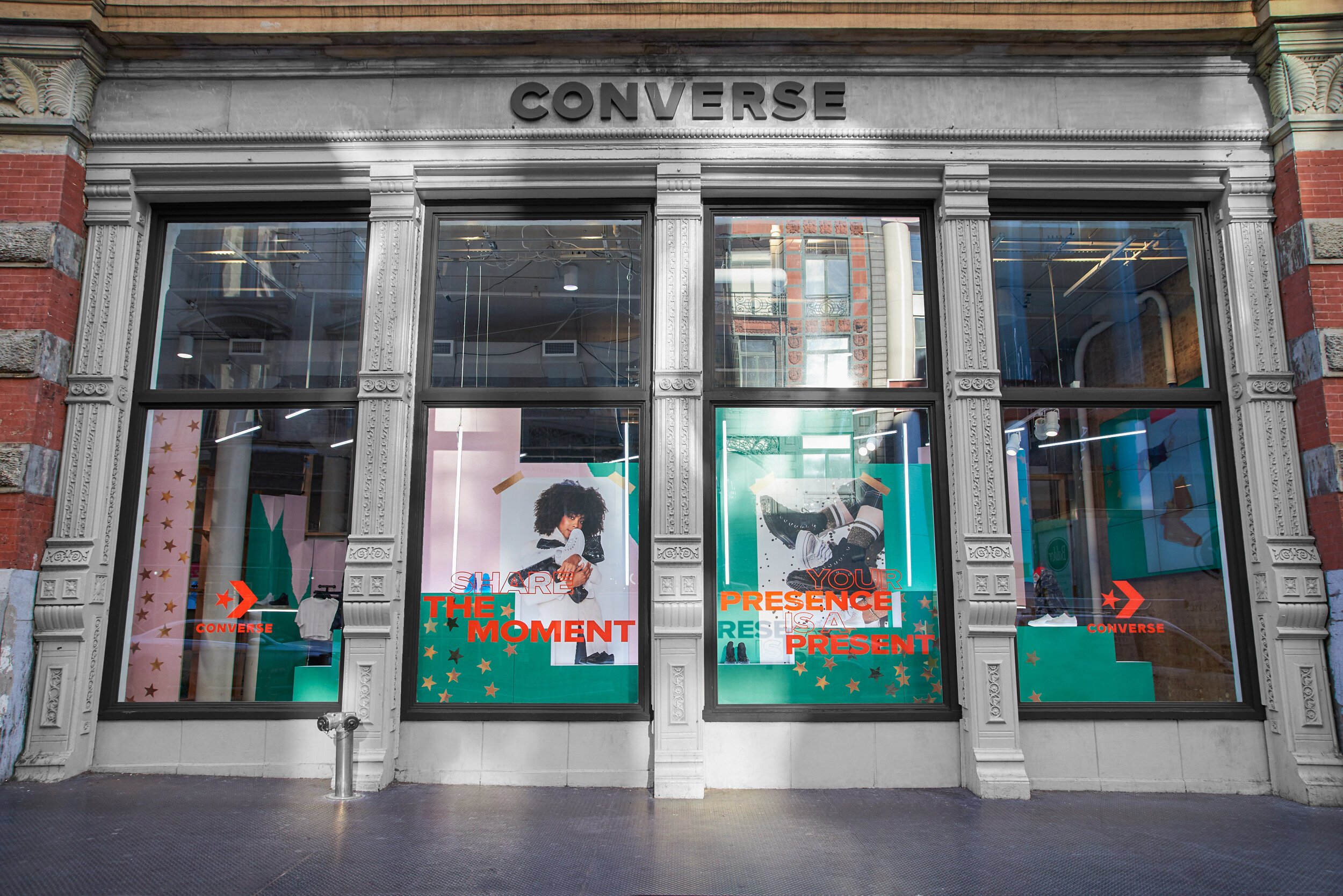 Outside view of a Converse store with large display windows, featuring posters and red text that reads 'Share the Moment' and 'Your Presence is a Present'.