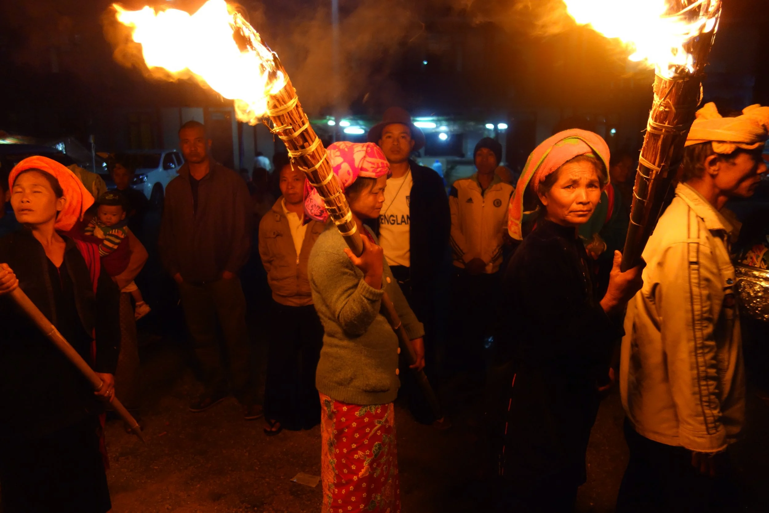 People participating in a night fire ritual, holding flaming torches, with some wearing colorful head coverings and traditional clothing, in an outdoor setting.