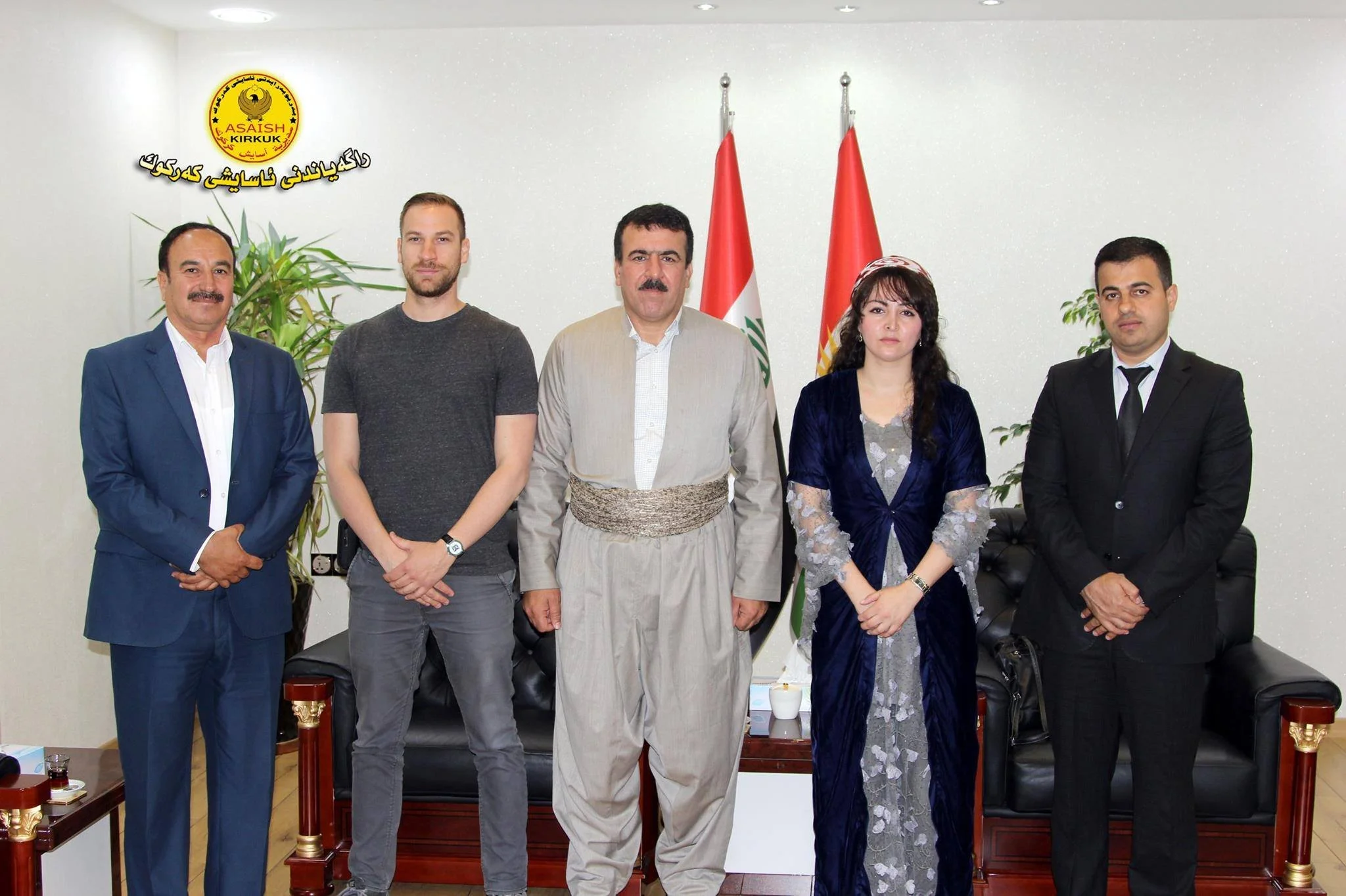Five people standing in a line in an office with Lebanese flags and a logo on the wall behind them. The group includes three men in suits, one man in traditional Middle Eastern attire, and one woman in traditional dress.
