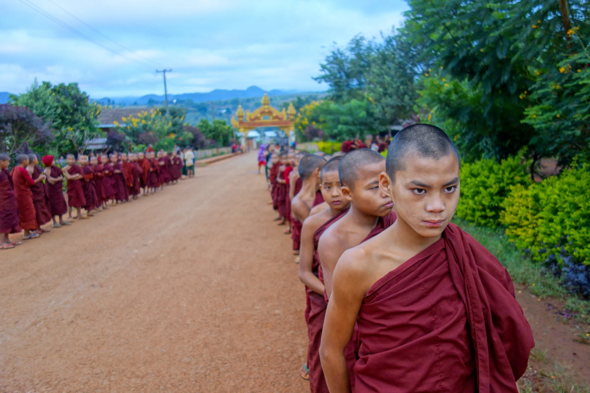 Line of young Buddhist monks with shaved heads wearing maroon robes walking on a dirt path in a rural area with lush greenery and a decorative gate in the background.