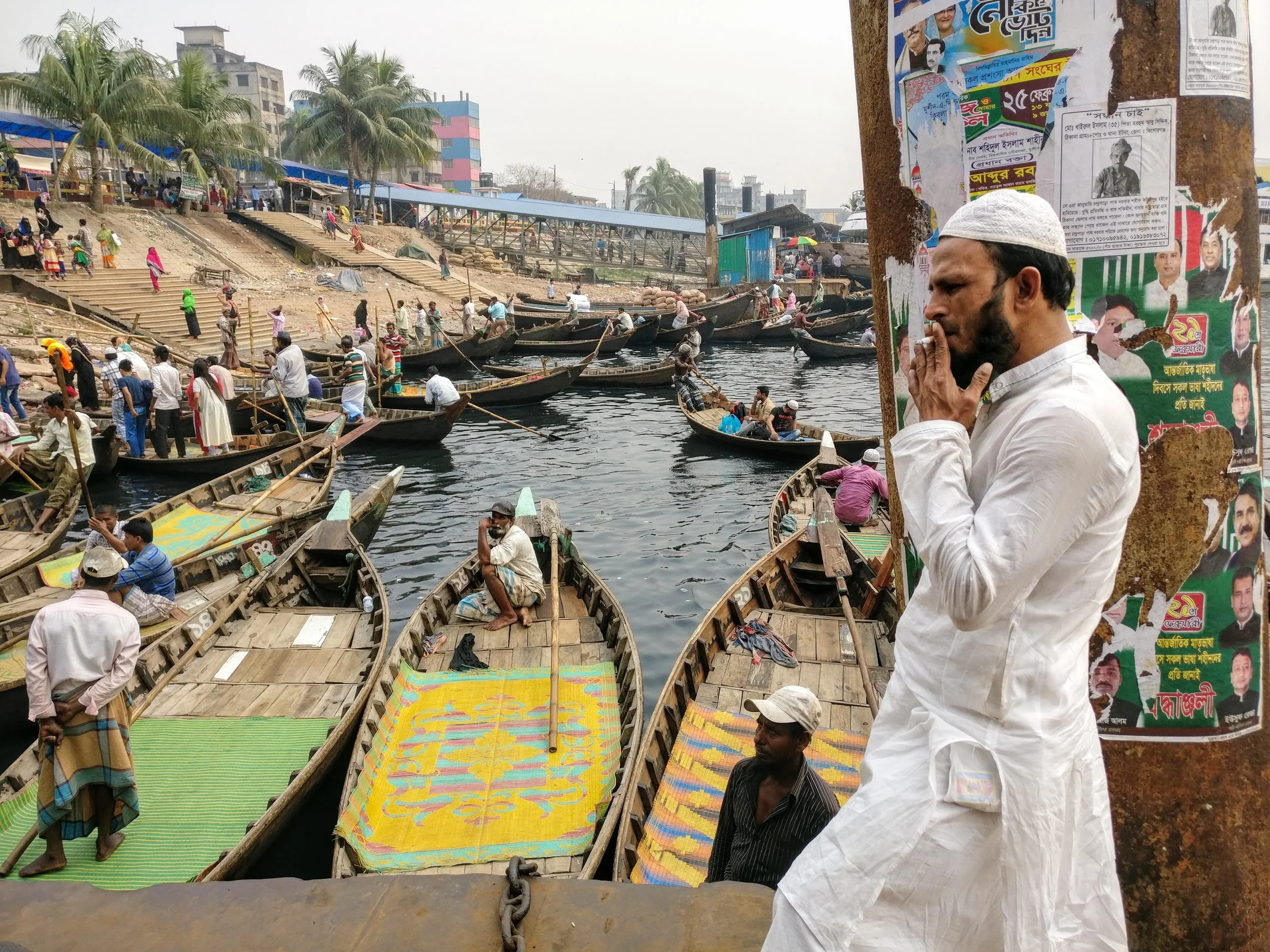A crowded dock area with numerous boats tied to the water, people walking and sitting around, some waiting in the boats. A man in traditional white attire with a white cap stands near a wooden post, appearing to be in deep thought. In the background,