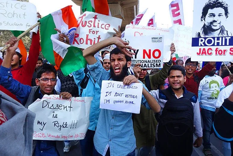Protesters holding signs and flags, advocating for justice for Rohit and opposing political issues, in an outdoor rally.