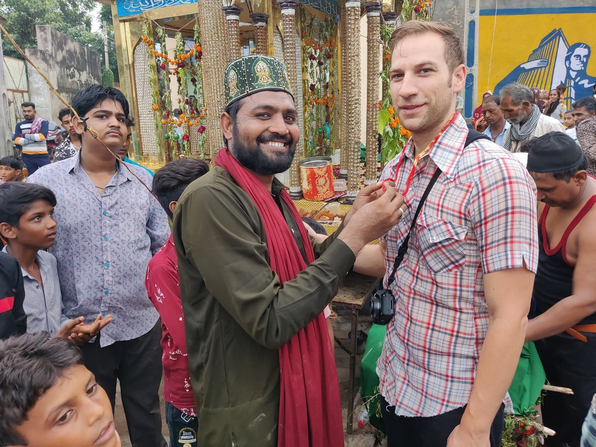 Two men, one in traditional attire with a cap and scarf, and the other in a plaid shirt, are smiling and engaging during a cultural or religious event.
