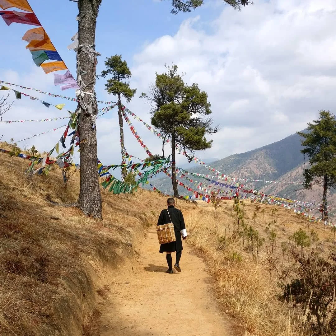 A person walking on a dirt path in Bhutan through a dry field with prayer flags strung between trees, mountains in the background, and a partly cloudy sky.