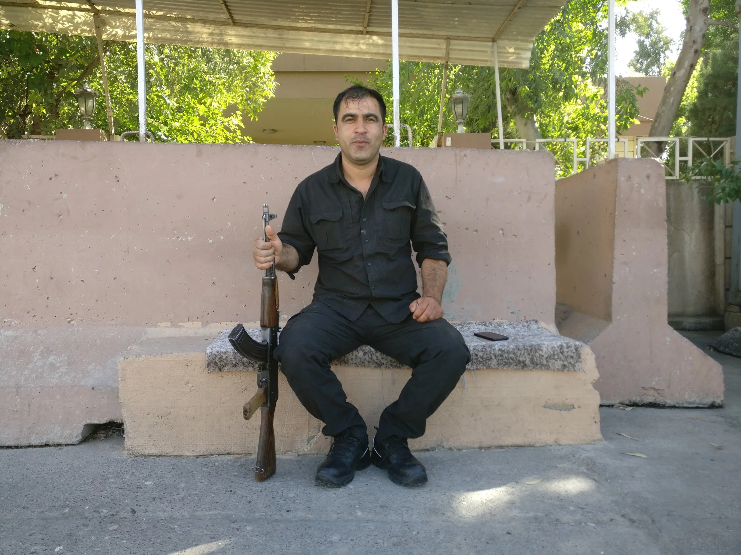 A man in black clothing sitting on a concrete bench outdoors, holding an AK-47 rifle, with a smartphone on the bench beside him.