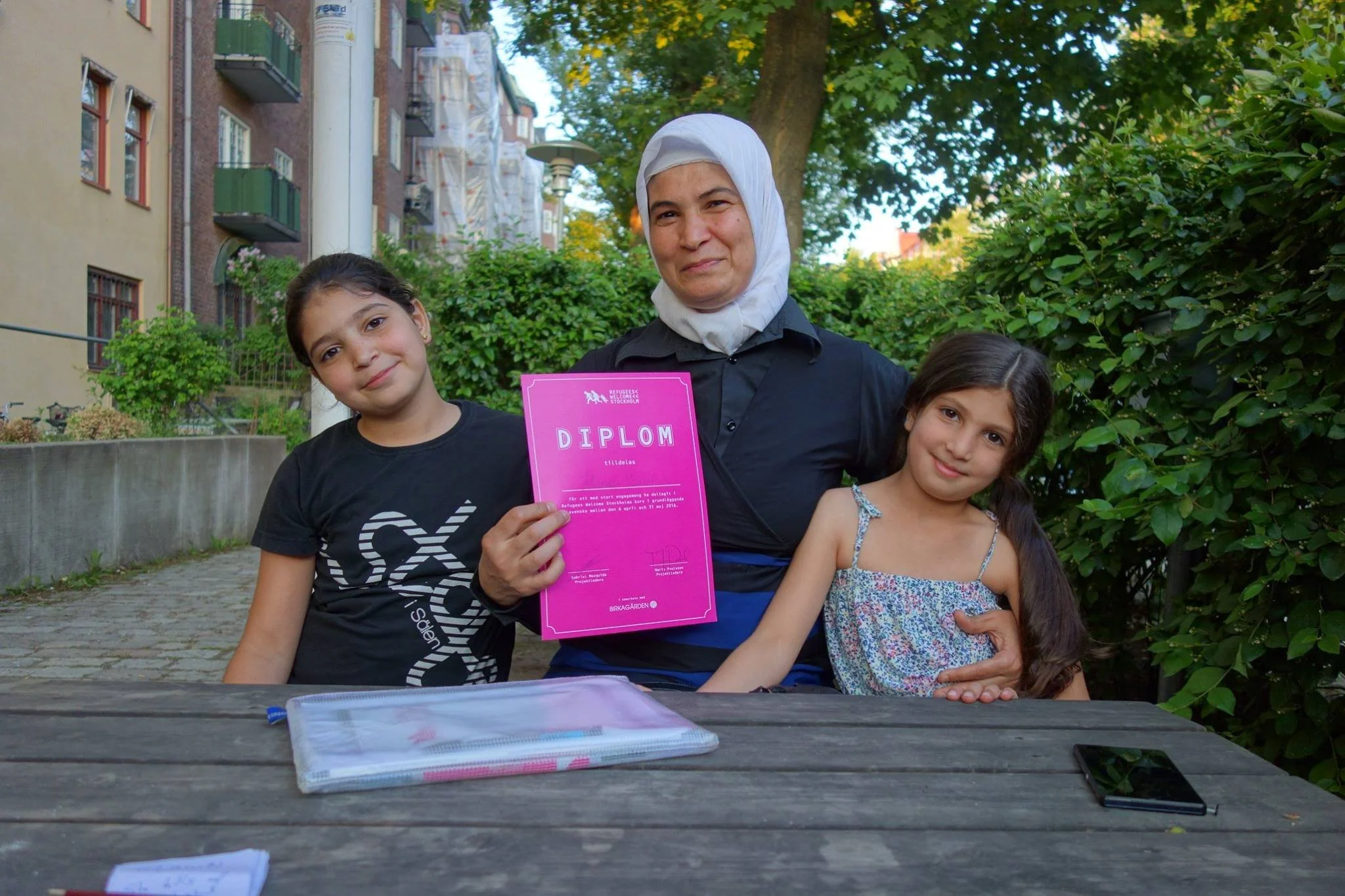 A woman wearing a white headscarf holds a pink diploma while sitting outdoors with two young girls, one on each side, at a wooden table in a park or garden area with trees and buildings in the background.