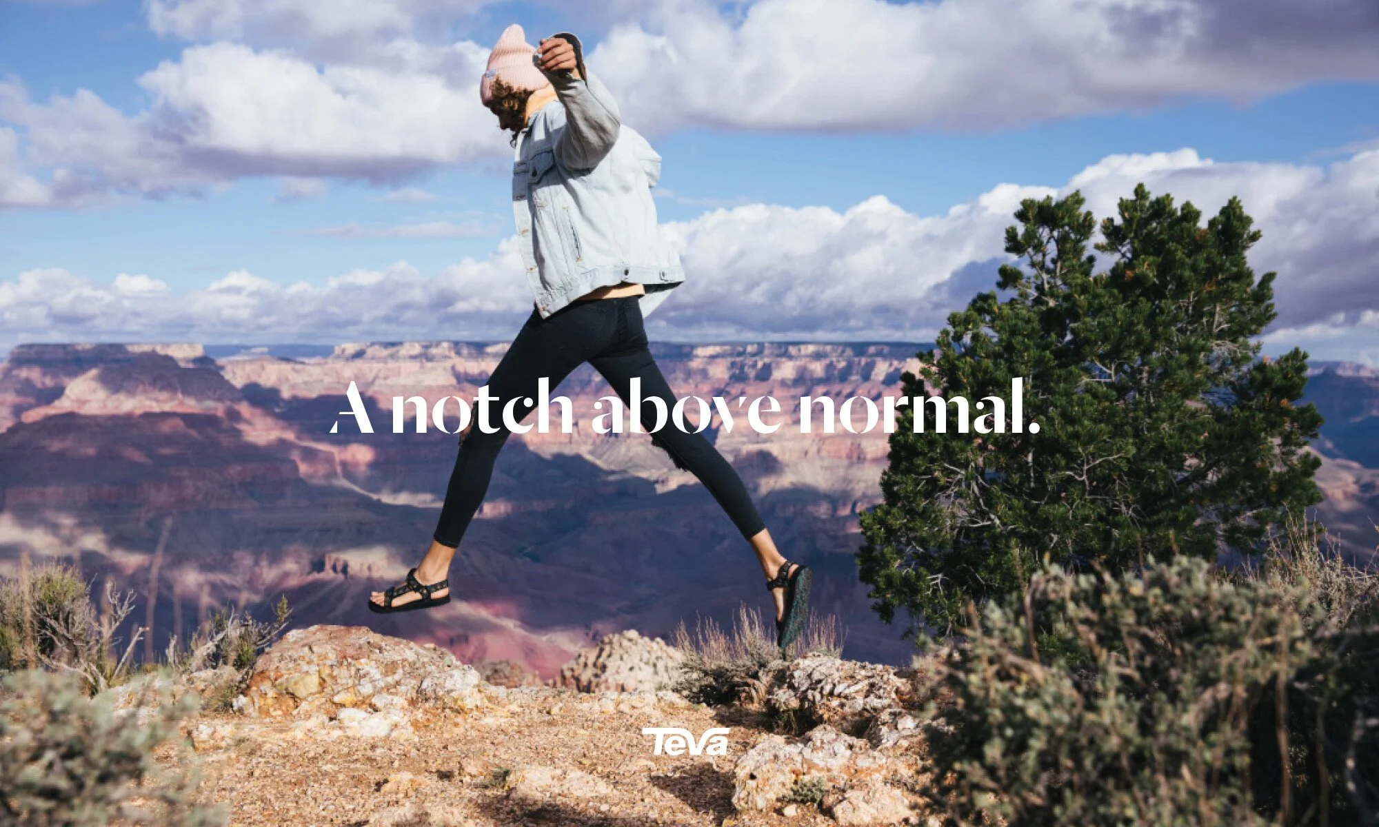 Young woman with curly hair jumping on rocks at the Grand Canyon, wearing a pink beanie, denim jacket, black leggings, and sandals, with a partly cloudy sky and a tree nearby.