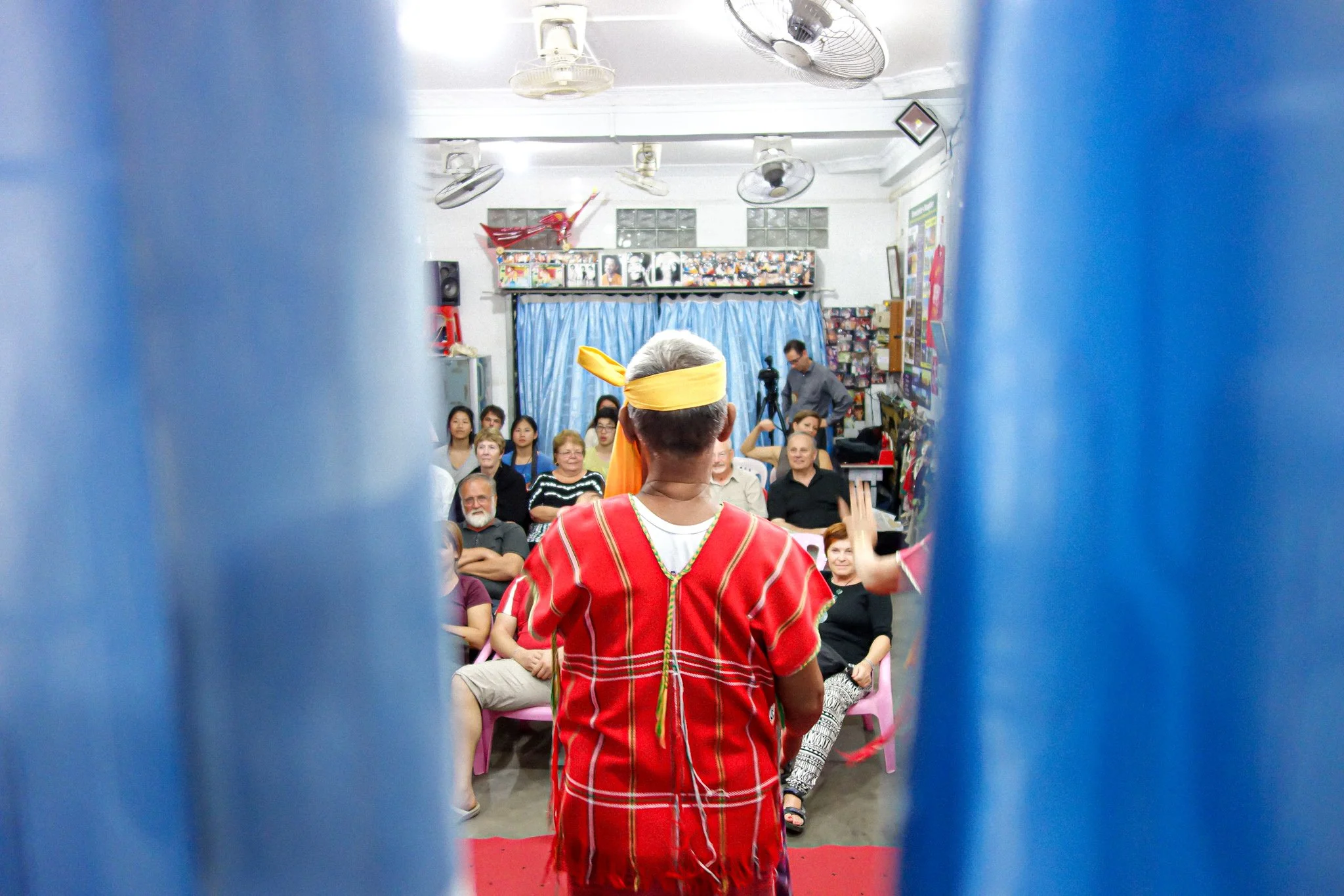 A performer in colorful traditional attire and a yellow headband faces an audience seated in a room with blue curtains, posters, and fans on the ceiling.