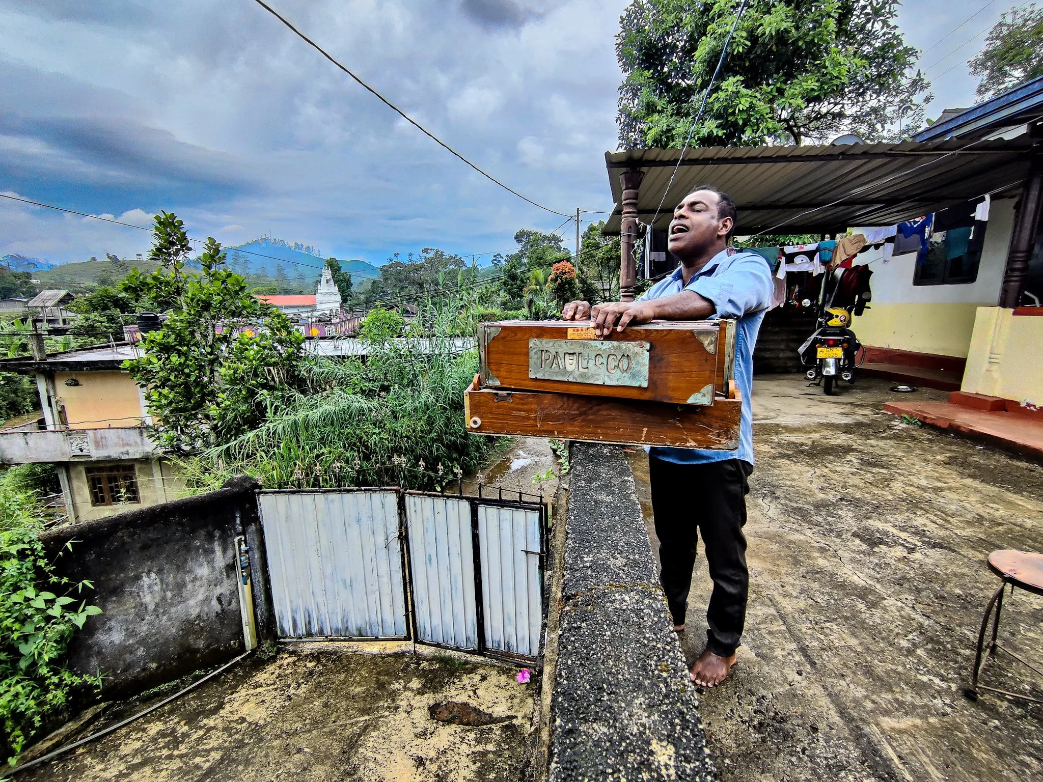 A man standing barefoot on a concrete balcony, singing with eyes closed in Hatton, Sri Lanka