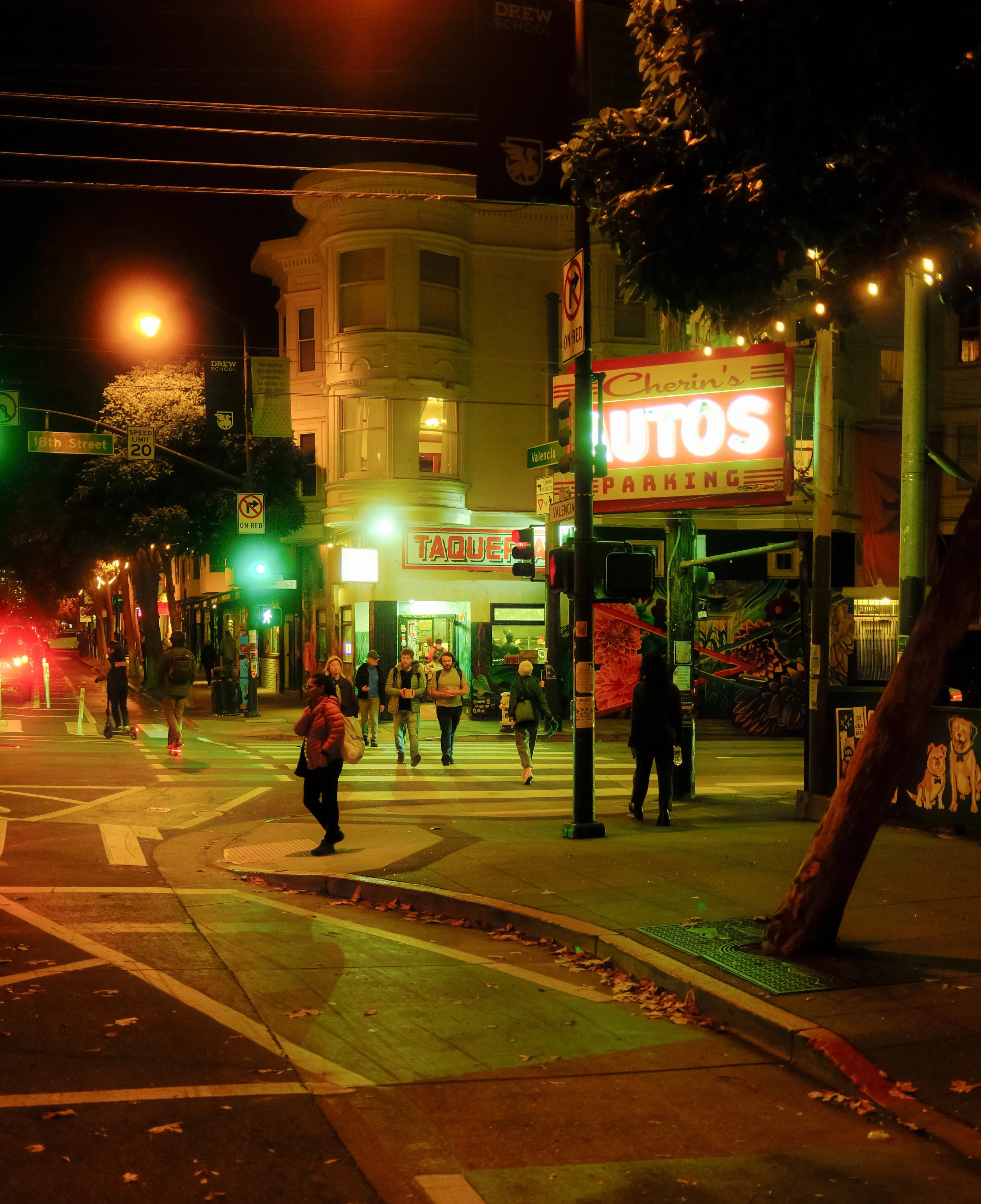 Nighttime street scene with people crossing at a crosswalk, illuminated signs for a taqueria and auto parking, and a corner building with bay windows. Streetlights and neon lights cast a warm glow.