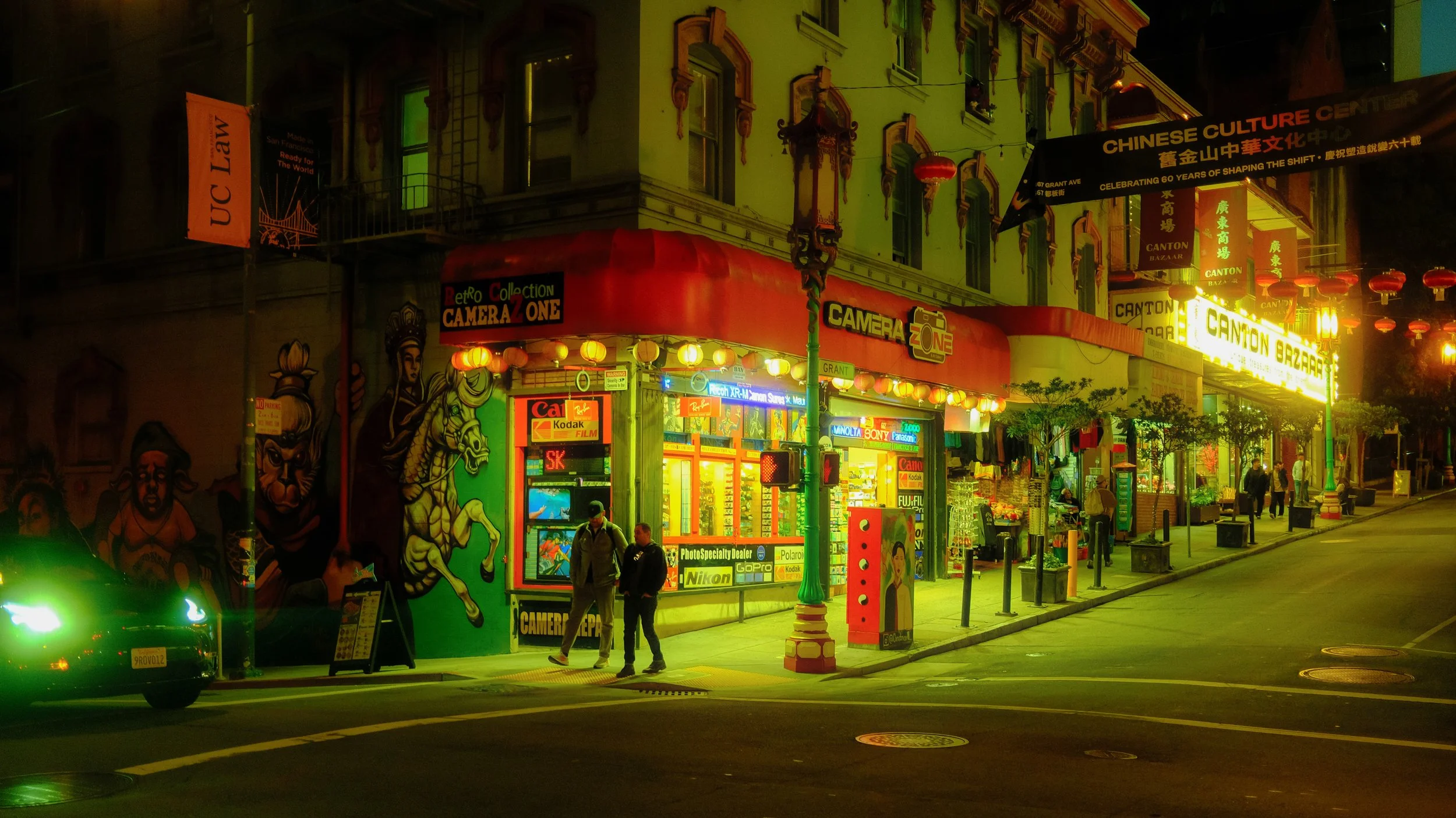 Night street scene in Chinatown with neon signs, a colorful storefront for cameras, murals of figures, and pedestrians walking.