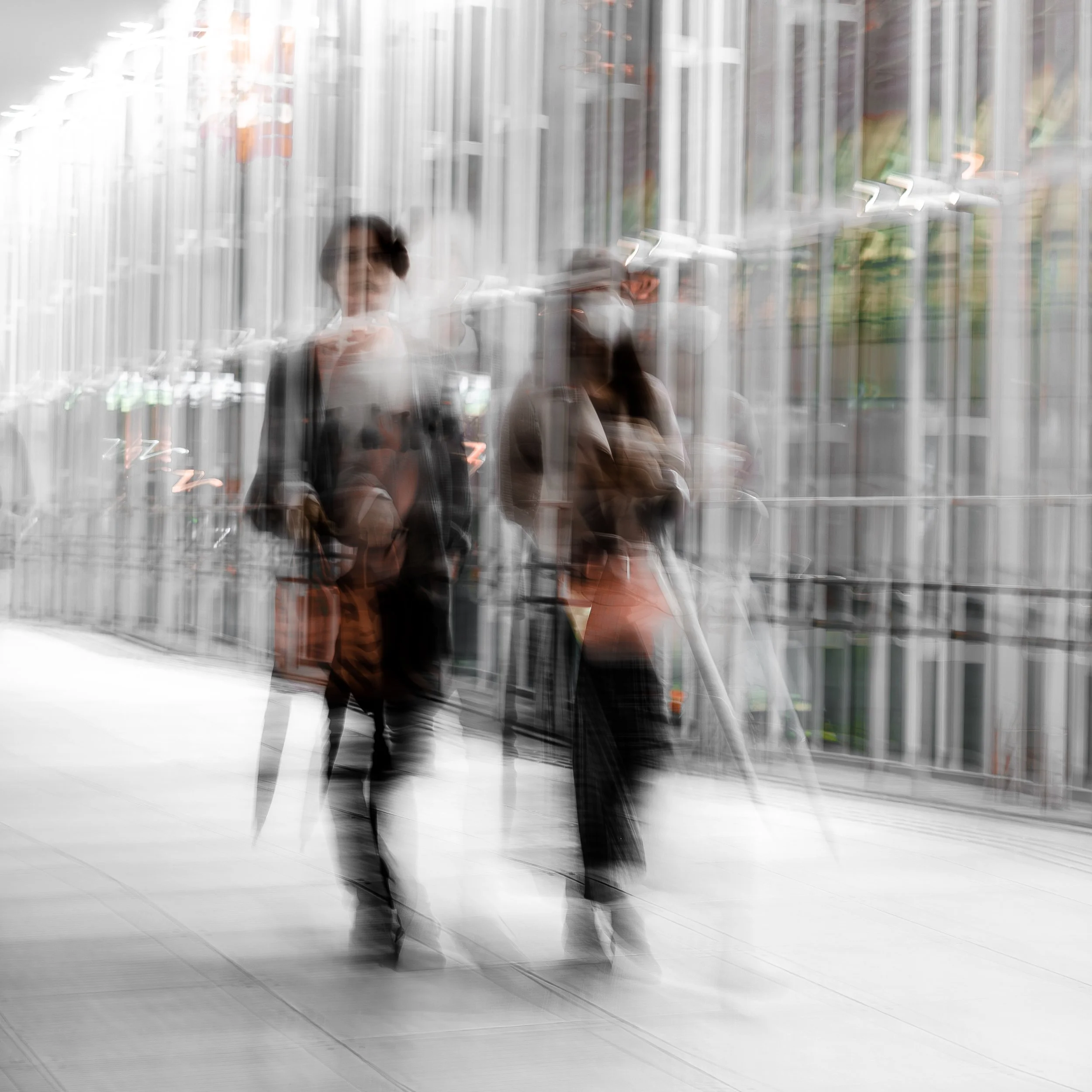 Blurry photo of two women walking outdoors in an urban setting, with glass buildings and trees in the background.