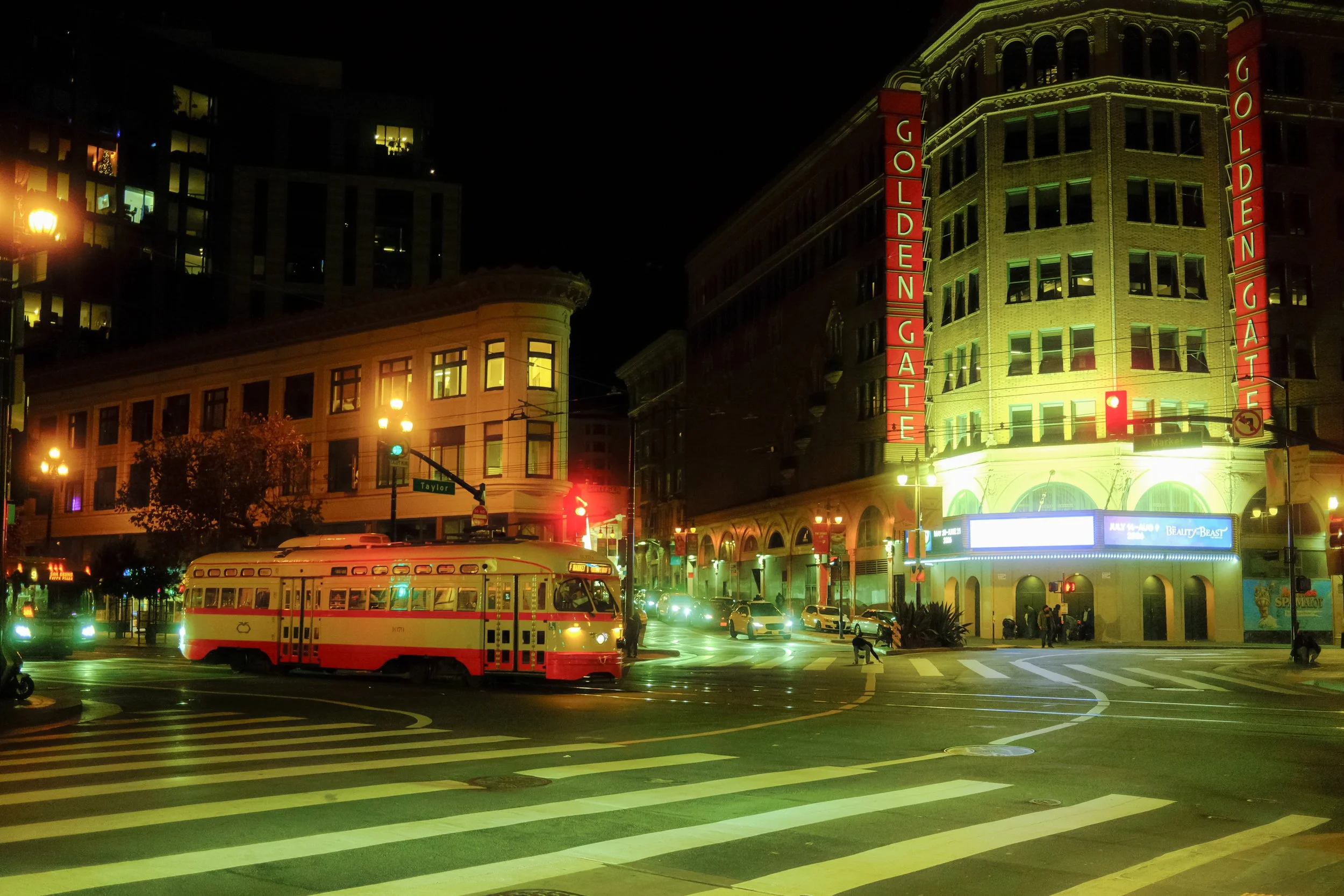 Night scene of a city intersection with a red and cream trolley bus, traffic lights, cars, pedestrians, and illuminated buildings, including the Golden Gate building with bright signage.