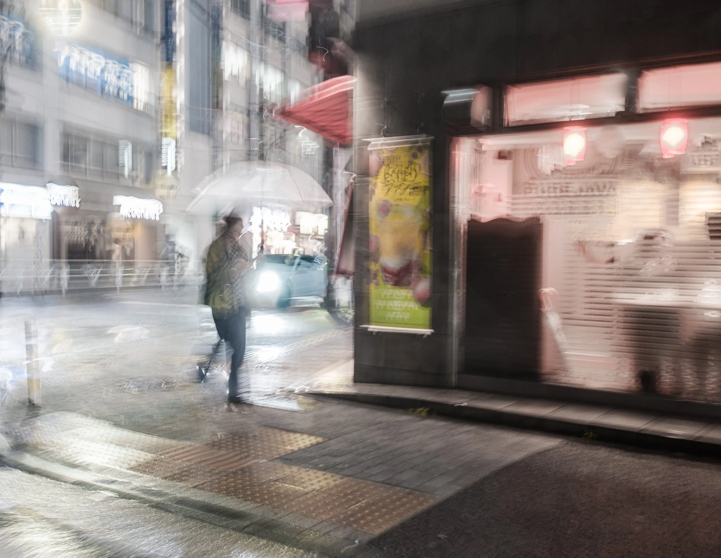 A person holding an umbrella walking on a rainy city sidewalk at night, with blurred storefronts and reflections on the wet pavement.