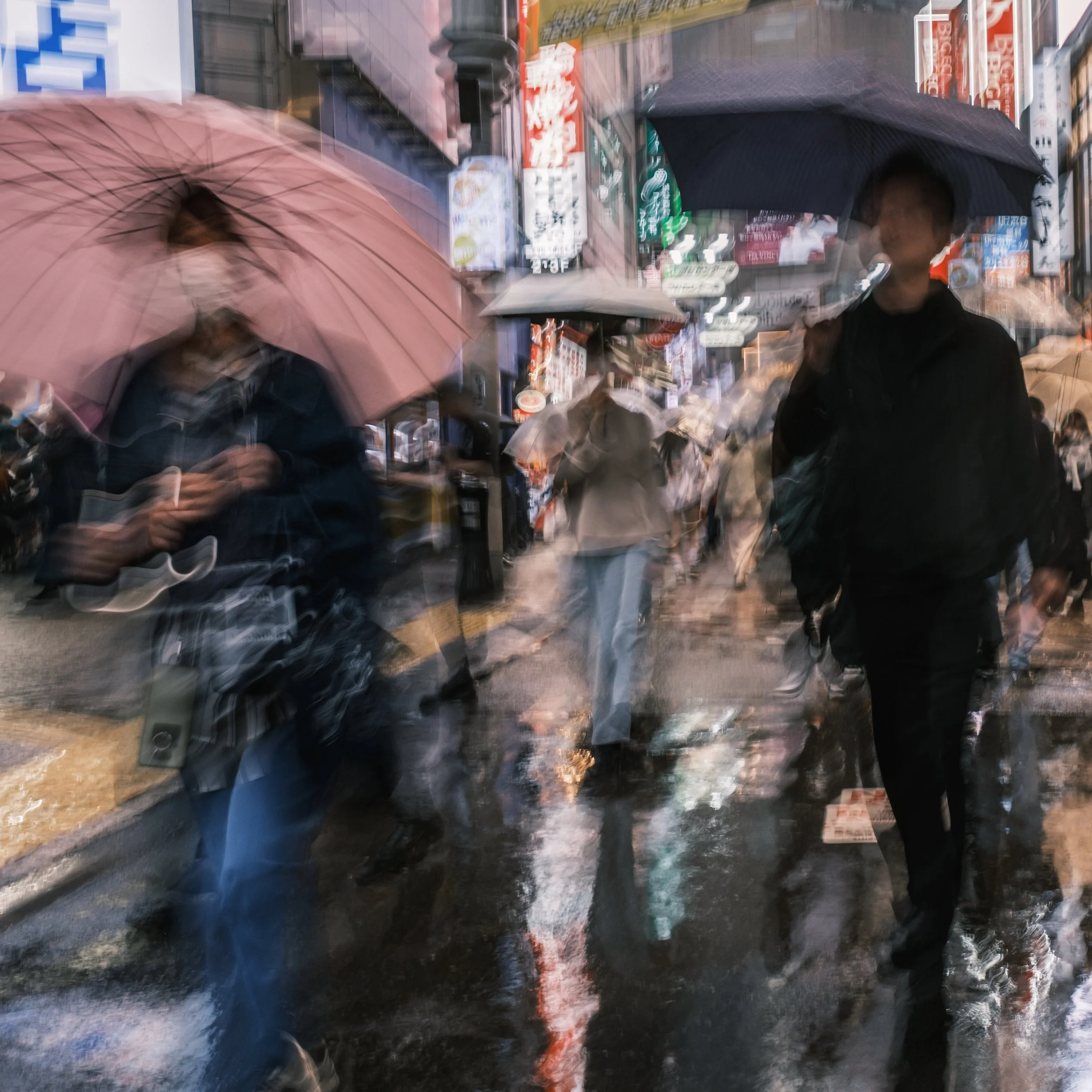 People walking with umbrellas in a busy, rainy street, with neon signs and storefronts in the background.