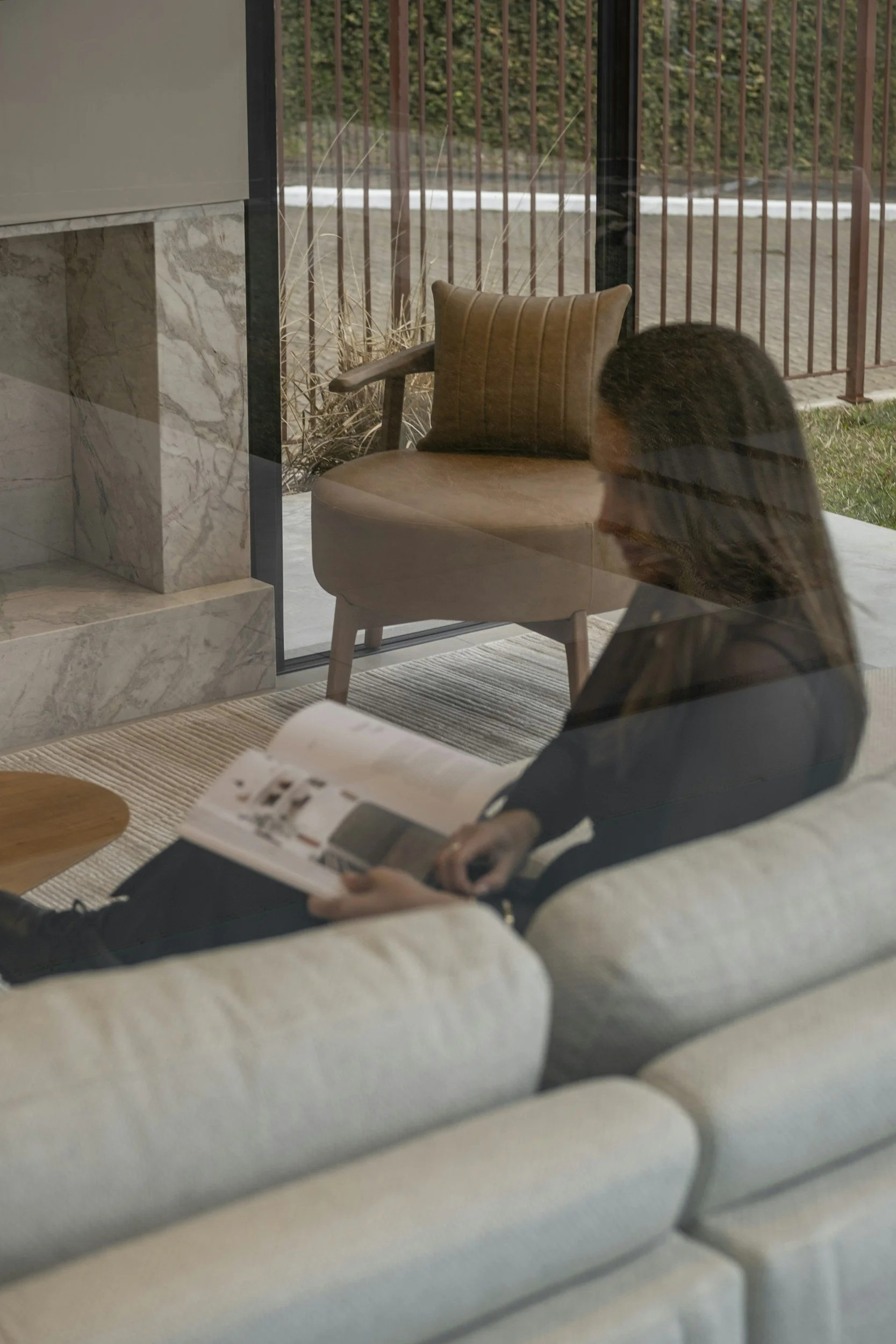 A woman sitting on a beige sofa reading a magazine, reflected in a glass wall, with a tan chair and outdoor scene visible through the glass.