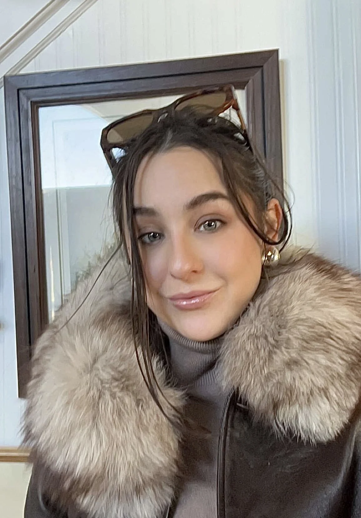 A young woman with brown hair, wearing a fur coat, earrings, and glasses on her head, posing in front of a mirror.