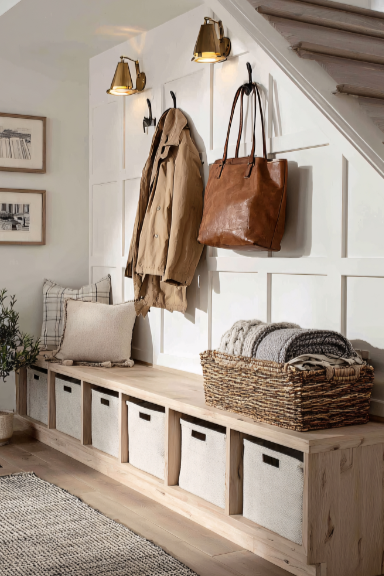 Entryway with coat hooks, a beige coat, a brown leather bag, wall-mounted lamps, and storage baskets under a wooden bench.