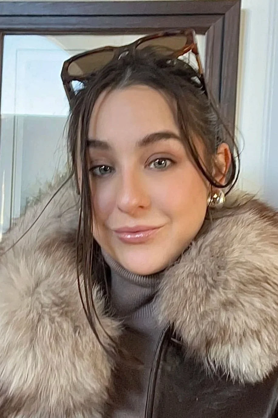 A woman with long brown hair, wearing sunglasses on her head, a fur-lined coat, and a light-colored turtleneck sweater, smiling at the camera.