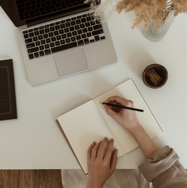 A person writing in an open notebook on a white desk with a laptop, a small bowl with clips, a tablet, and a vase with dried flowers.