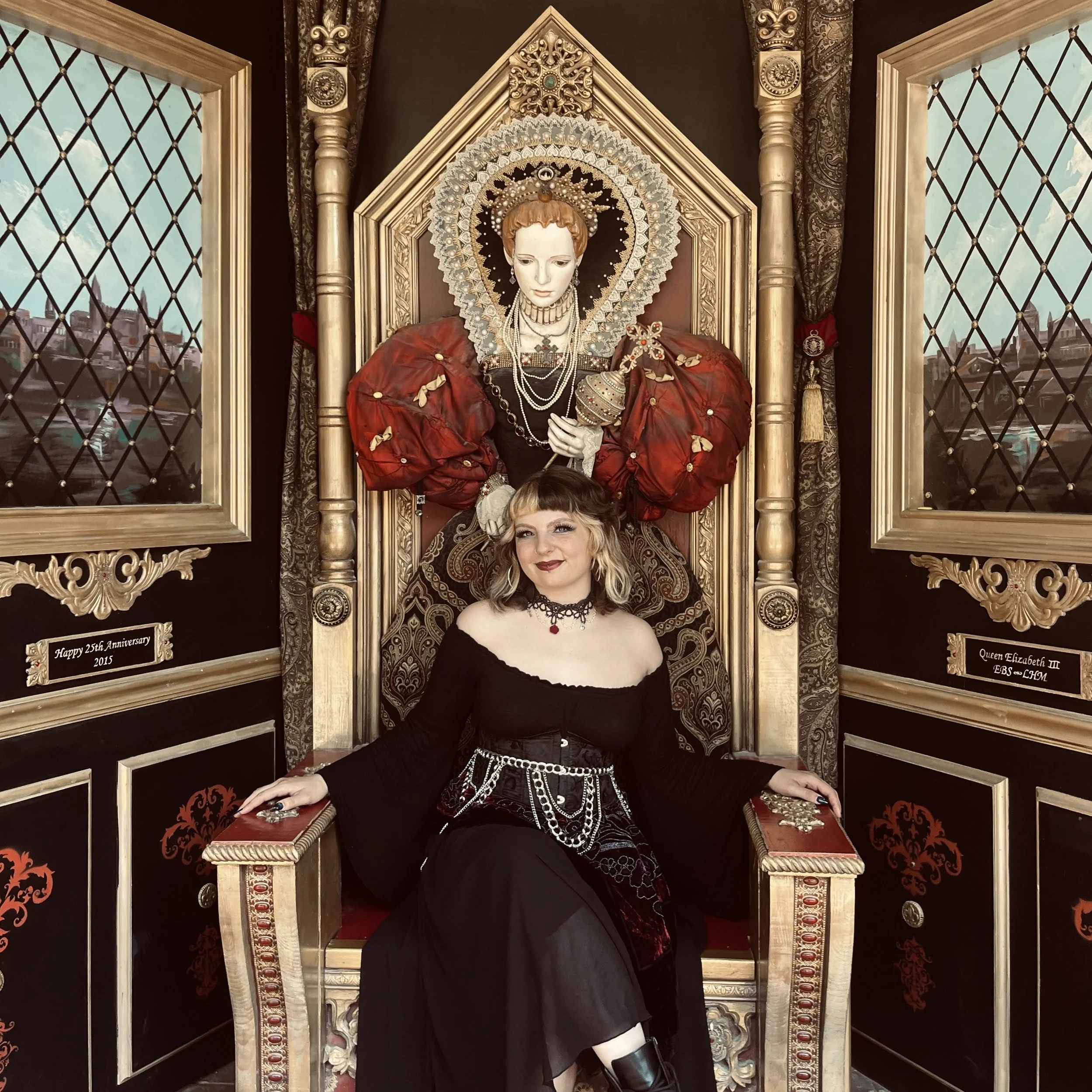 A young woman with blonde hair, wearing a black off-shoulder dress and gothic jewelry, sits on a decorated throne-like chair in a richly decorated room. Behind her is a regal statue of a royal woman with red hair, ornate jewelry, and a crown-like headpiece, set within an elaborate gilded frame.