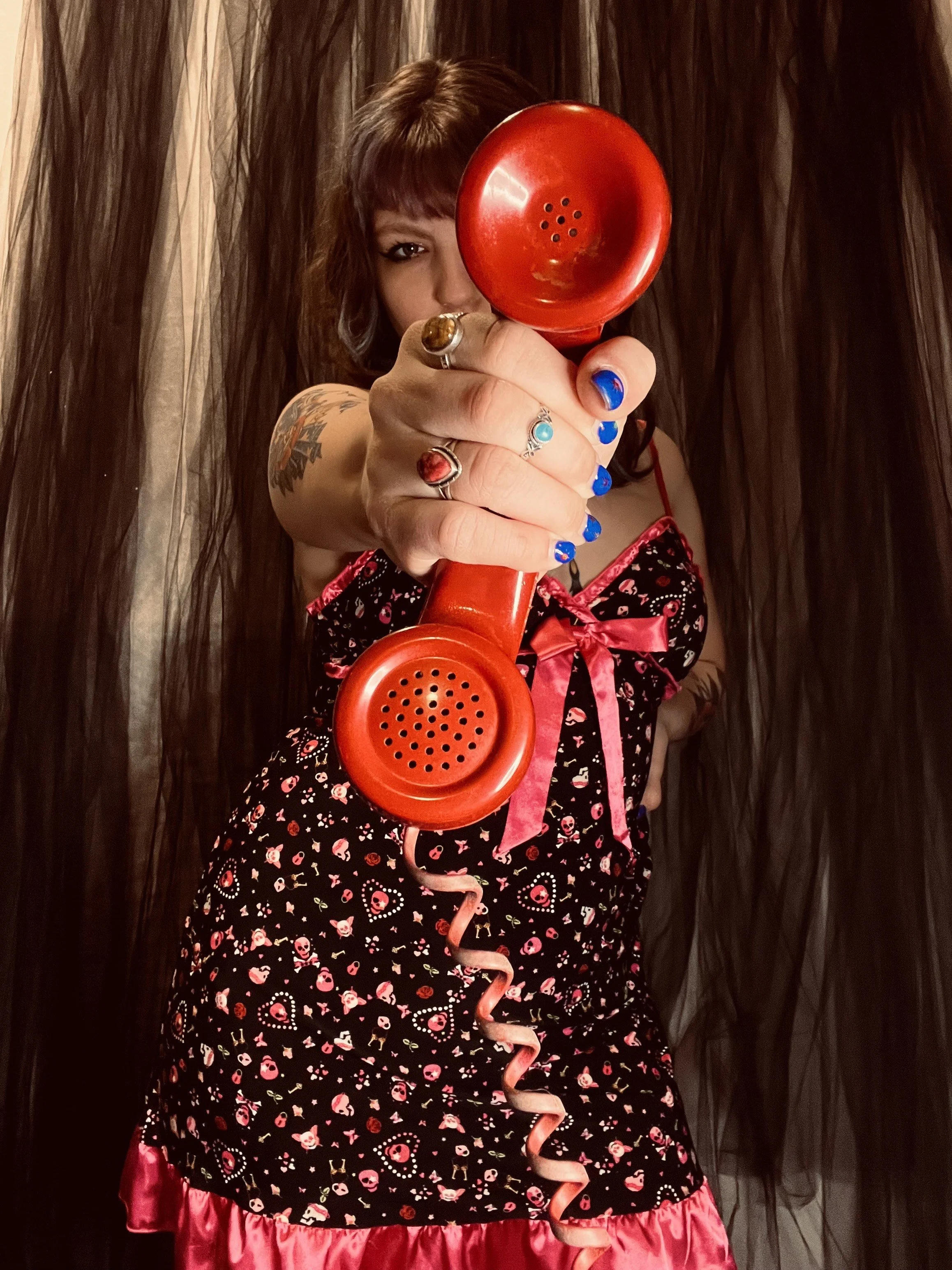 A woman with dark hair and tattoos on her arm, wearing a black dress with pink skull and heart patterns and pink trim, holding a red vintage telephone receiver toward the camera, with a black curtain backdrop.