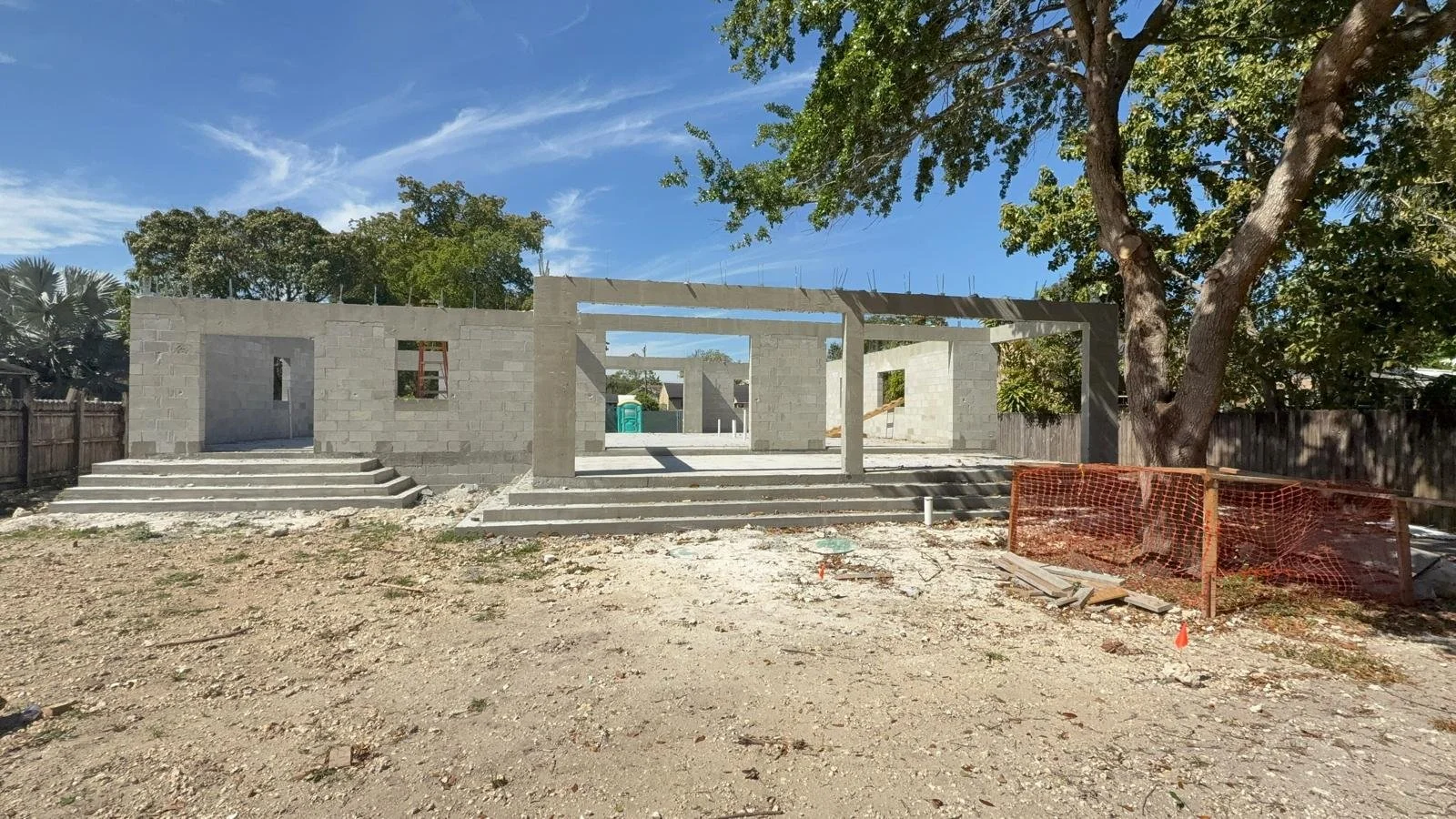 Construction site with a partly built concrete structure, stairs leading up to the entrance, and a large tree on the right side, next to an orange safety fence, under a clear blue sky.
