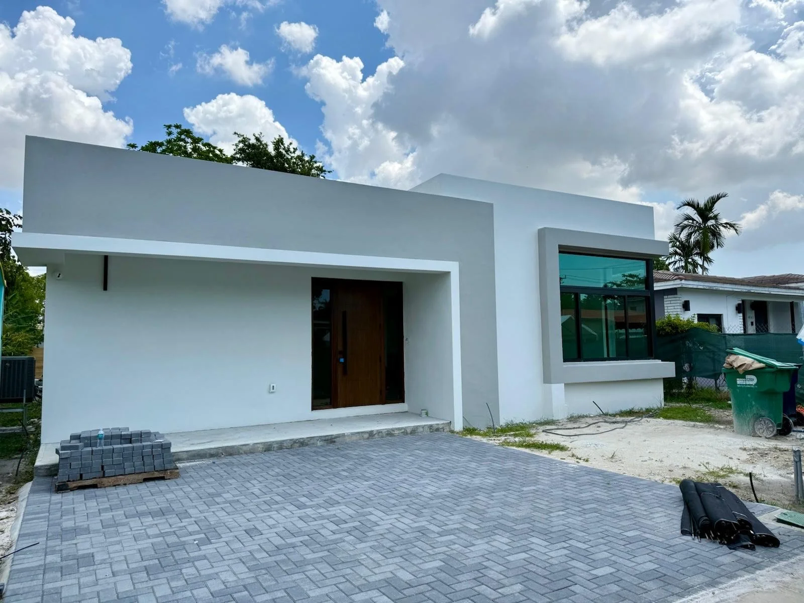 Modern white house under construction with a paved driveway, a large window, and a wooden front door, surrounded by construction materials and equipment.