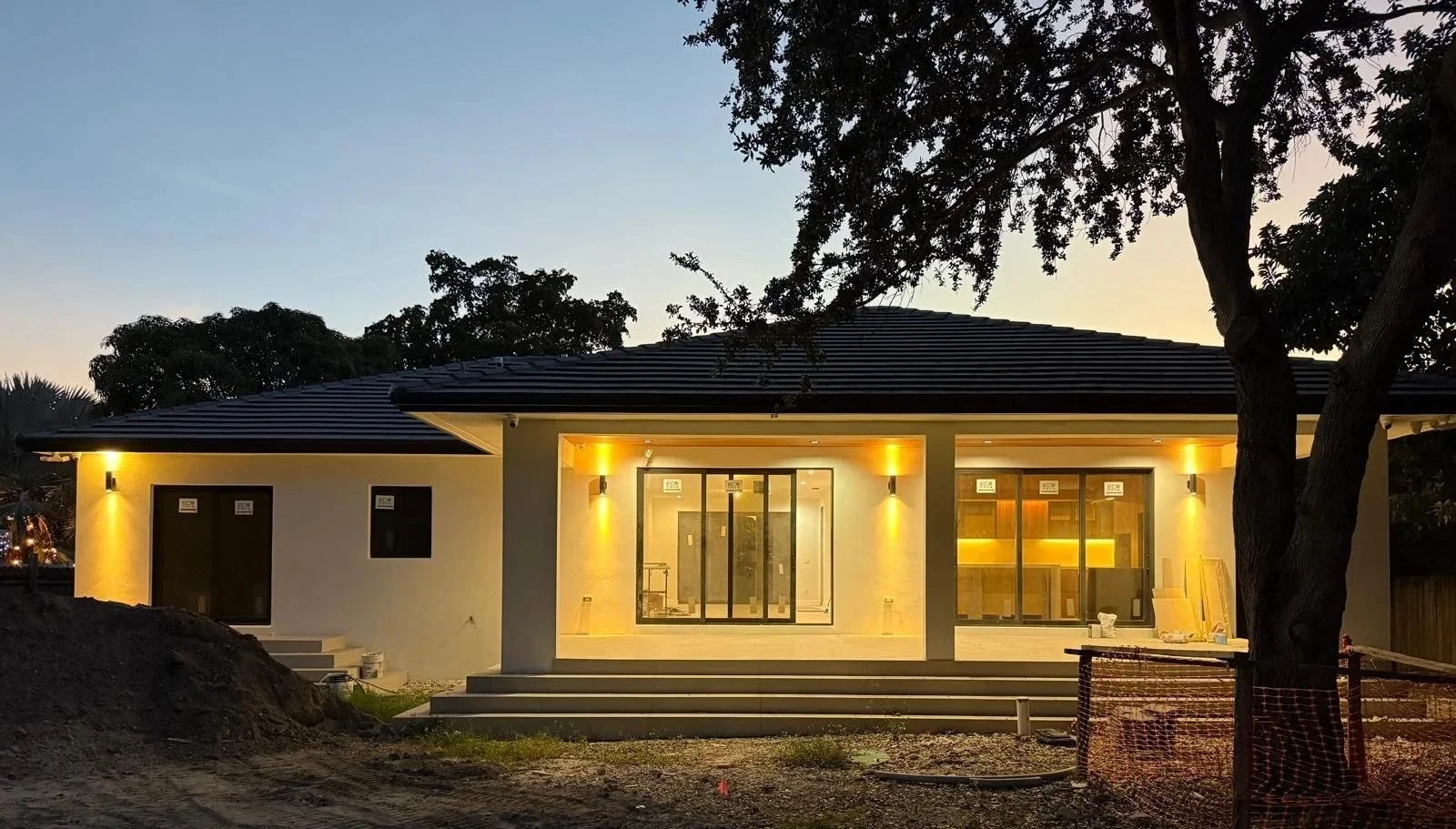 A house under construction with exterior lighting, steps leading up to the entrance, and windows with interior lighting, surrounded by trees at dusk.