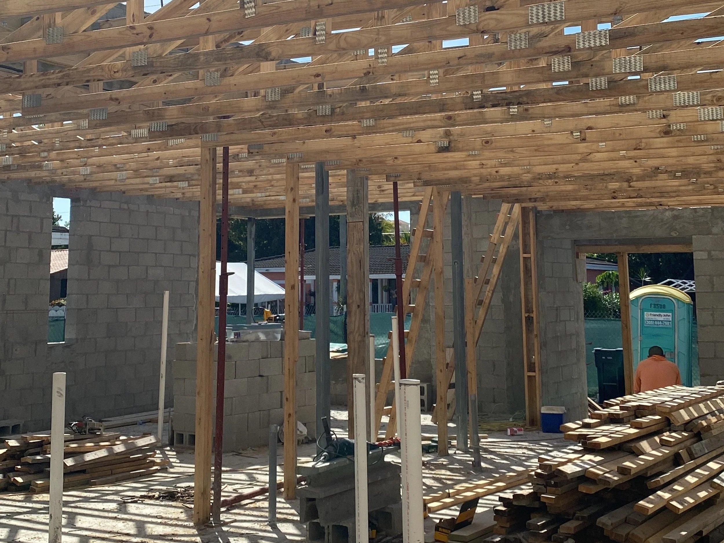 Construction site with wooden beams and scaffolding, worker in orange shirt near porta-potty, stacks of lumber, incomplete brick walls.