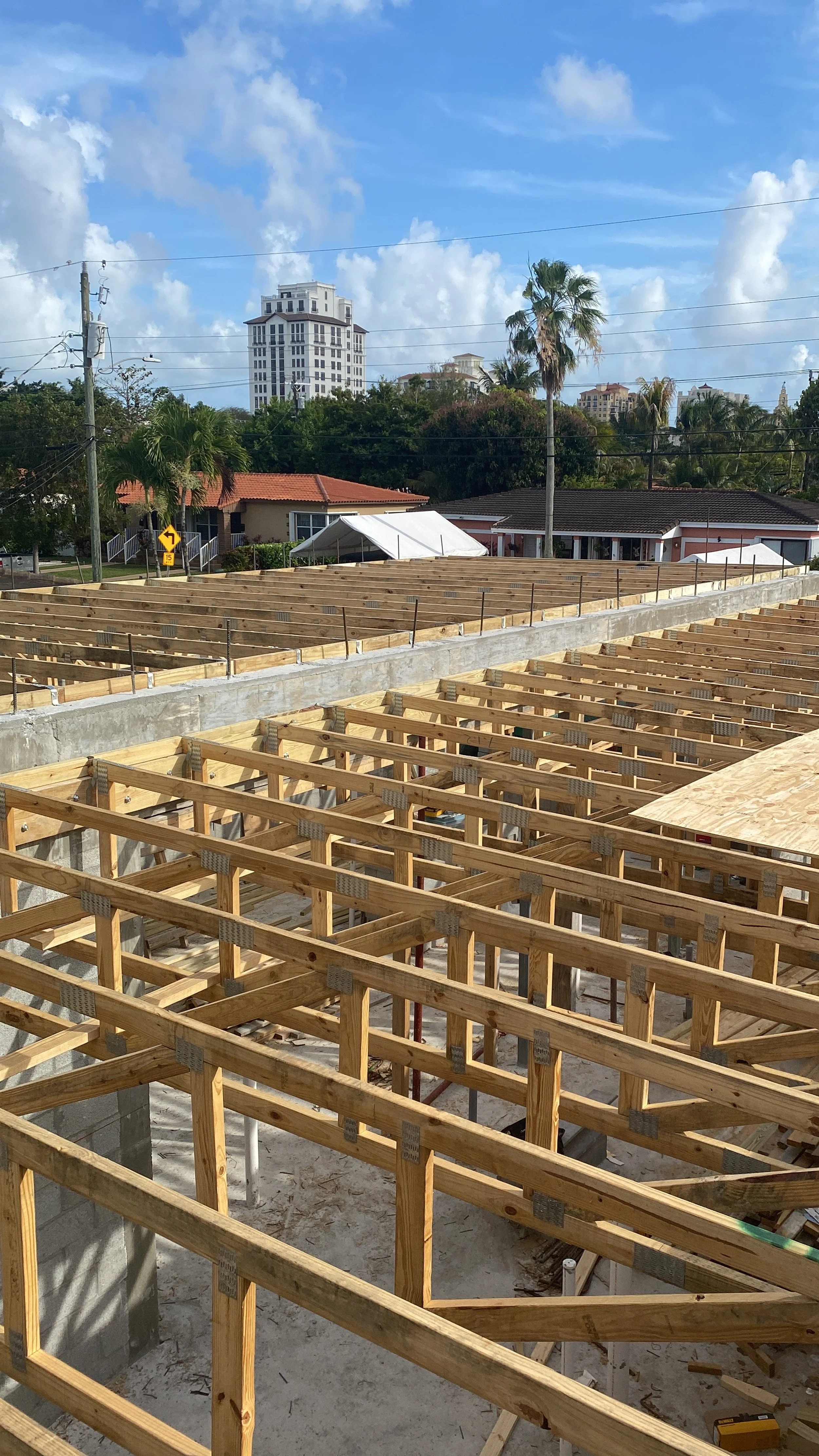 Construction site with wooden framing for a building, against a backdrop of trees, houses, and a tall apartment building under a partly cloudy sky.