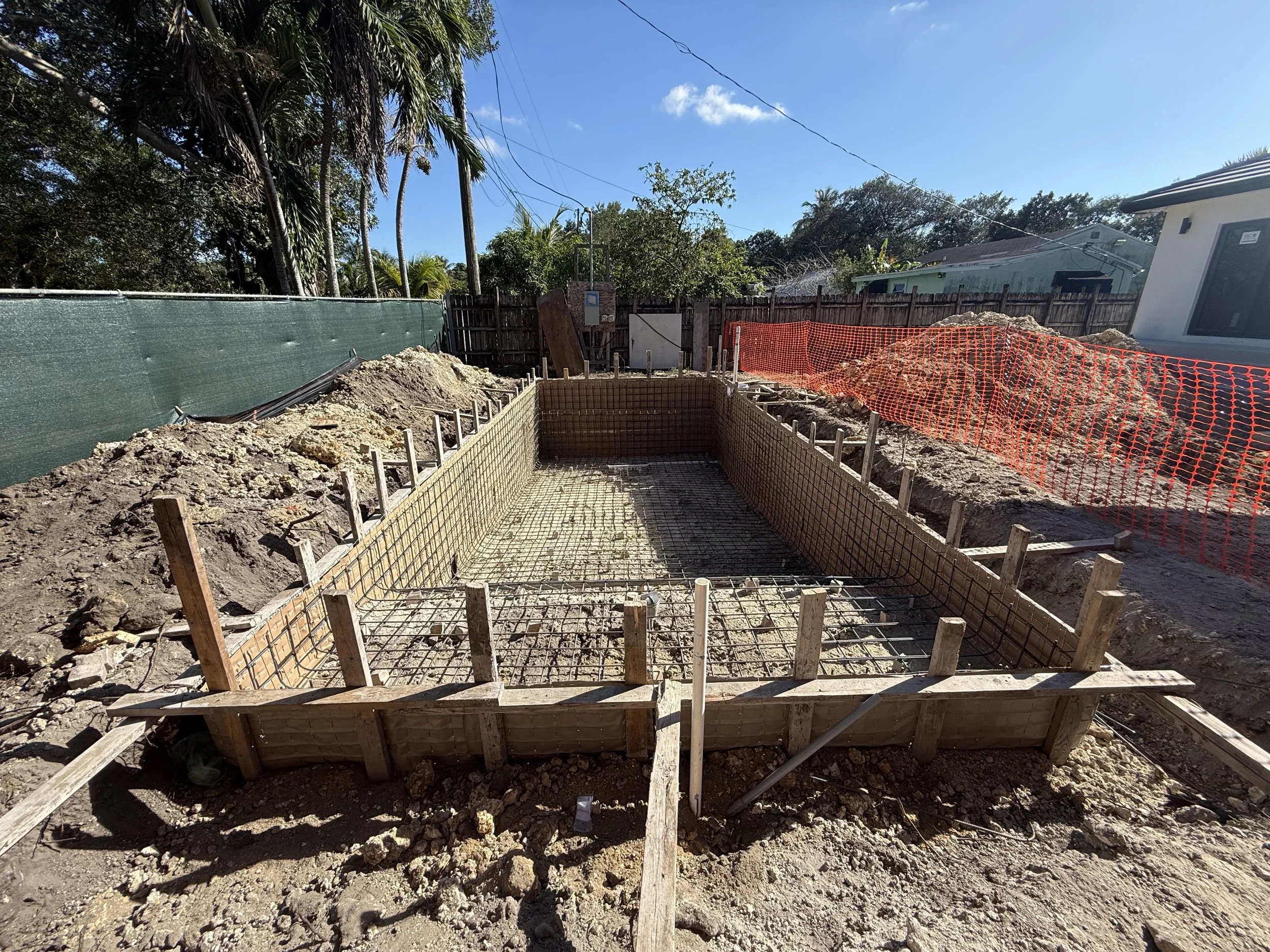 Construction site with excavated area framed by wooden barriers, rebar mesh inside, orange safety fencing along the back, and trees and houses in the background.