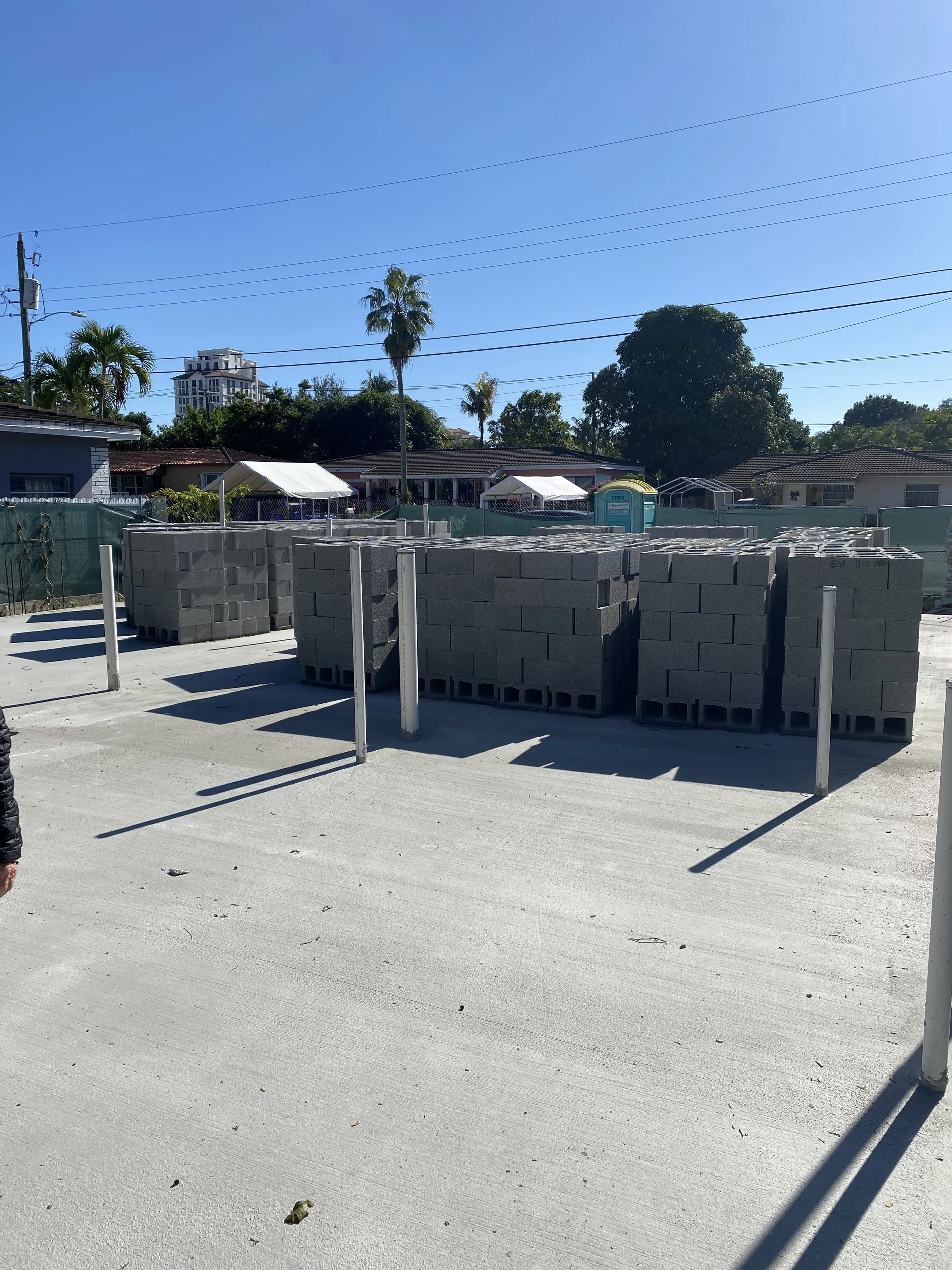 Construction site with stacks of concrete cinder blocks and metal posts on a concrete surface, with a background of trees, houses, and a clear blue sky.