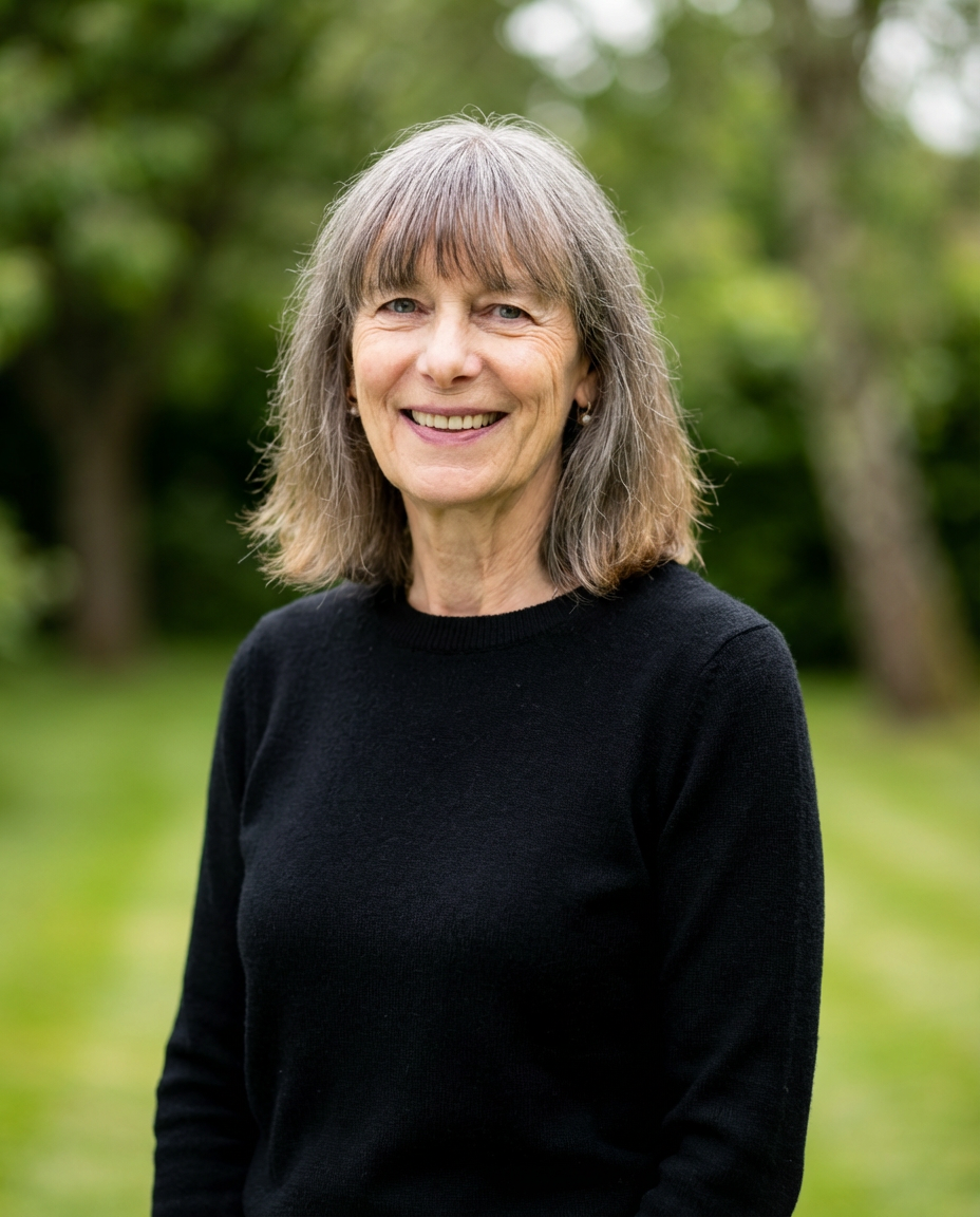 An older woman with shoulder-length gray hair, smiling at the camera, wearing a black sweater outdoors with trees and greenery in the background.