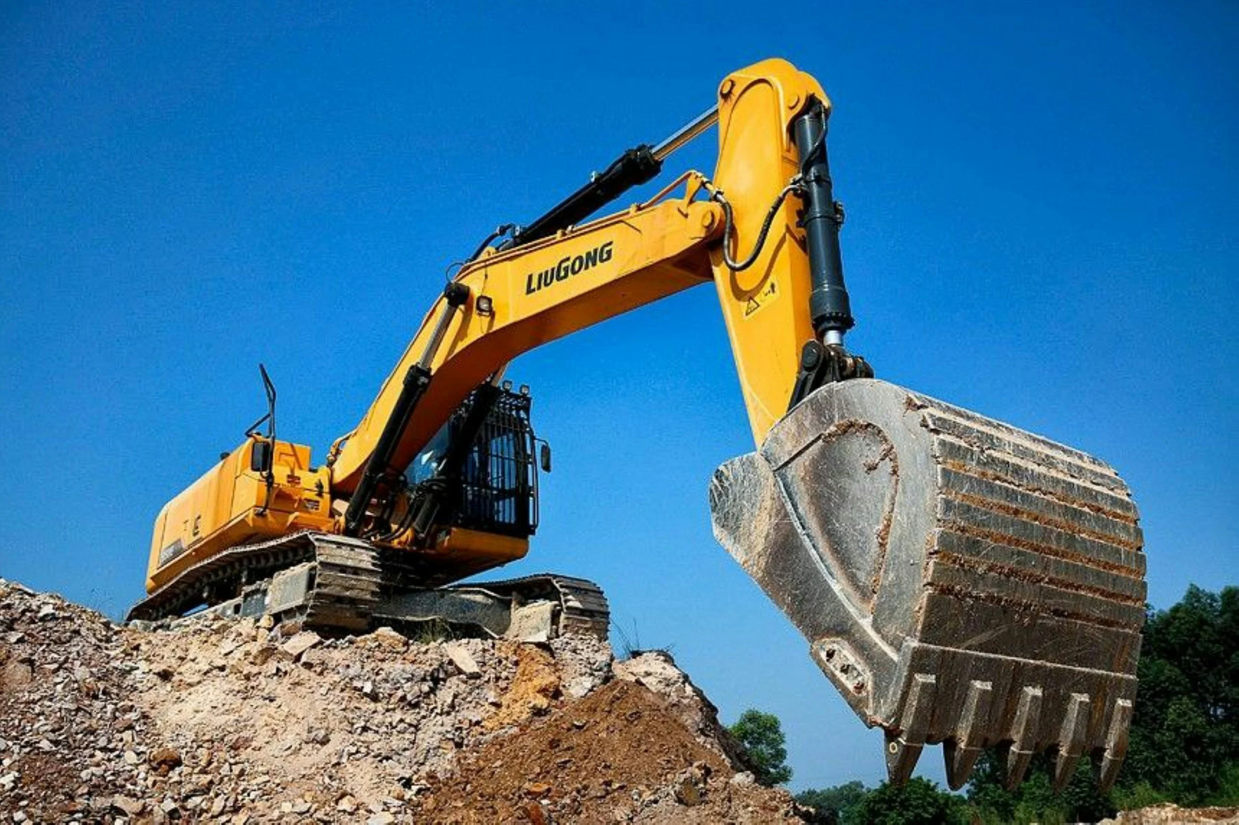 Yellow excavator with 'LIUGONG' logo on a blue sky background, digging into dirt and rocks with its large metal bucket.
