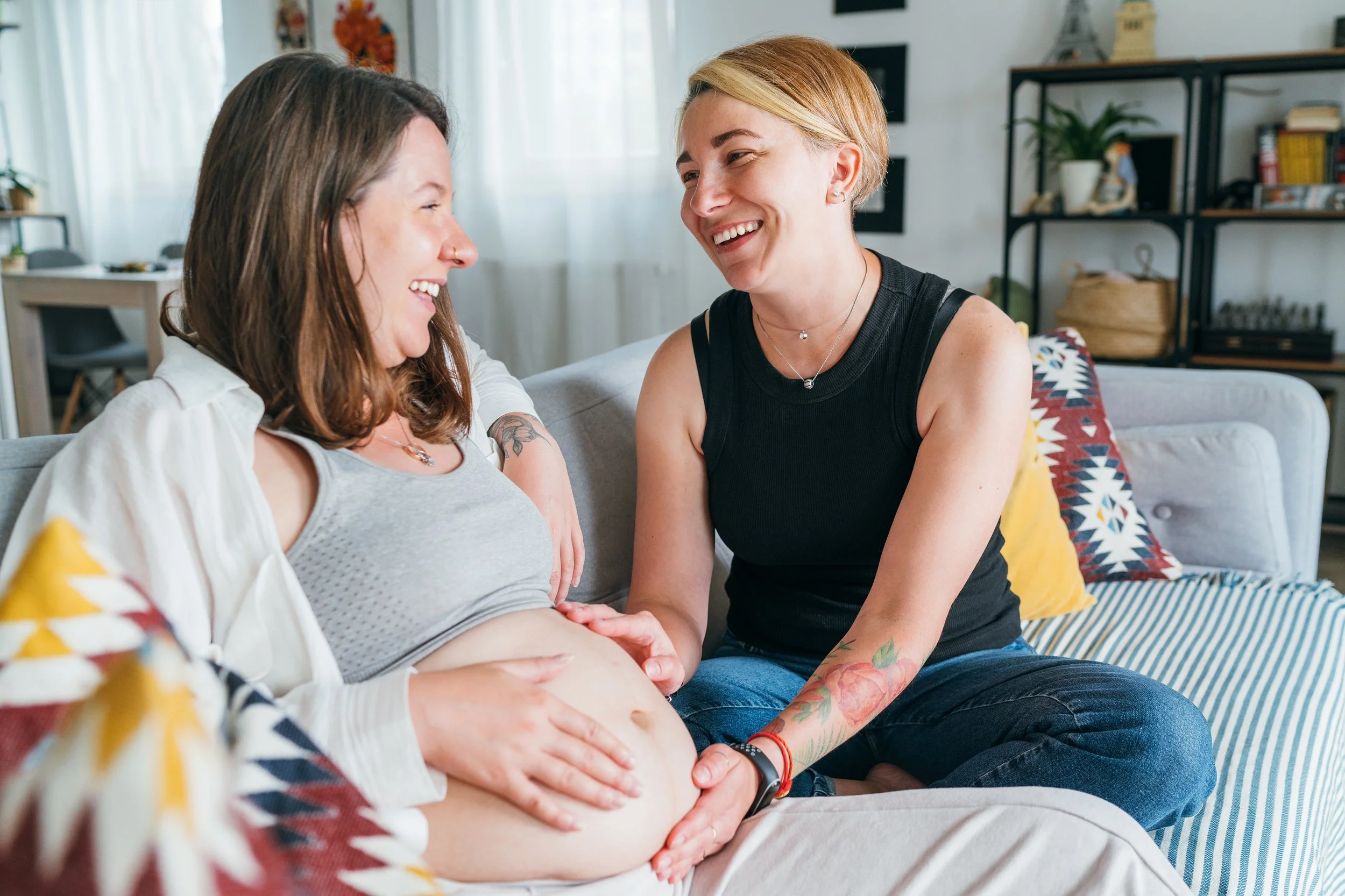 Two women sitting on a couch, one pregnant and smiling, the other touching her belly, in a cozy living room with cushions and shelves in the background.