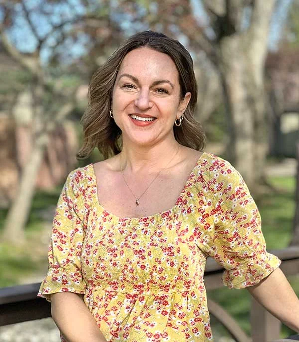A woman with shoulder-length brown hair, smiling, wearing a yellow floral dress and small earrings, standing outdoors in a park with trees in the background.