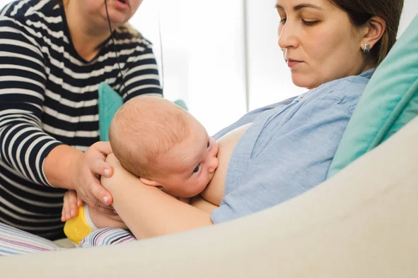 A woman in a hospital bed with a newborn baby on her chest, while a person in a striped shirt assists nearby.