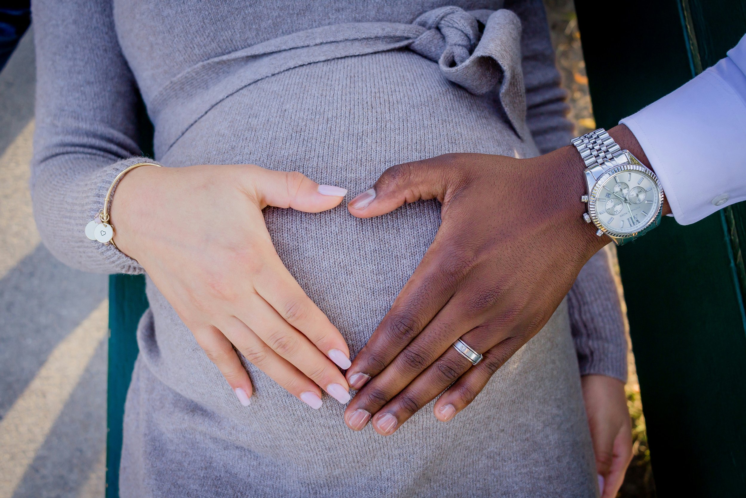 Close-up of a pregnant woman and partner forming a heart shape with their hands on her belly, sitting on a bench outdoors.