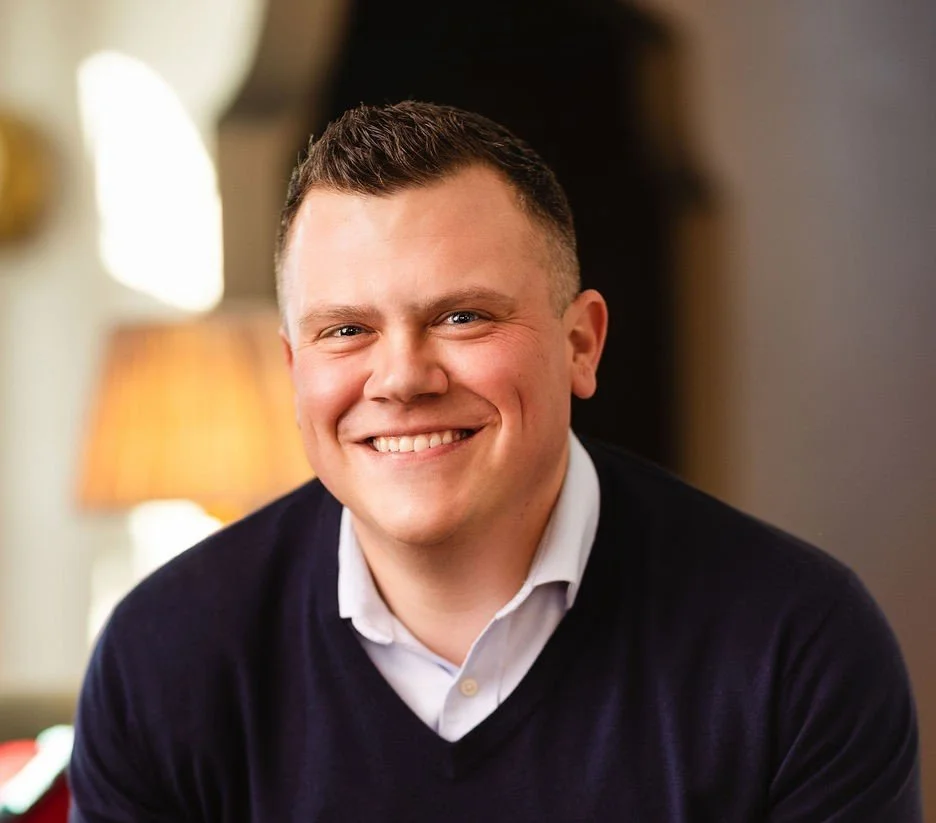 Justin Solomon, a young man with short hair smiling in a warmly lit room, wearing a white collared shirt and a dark sweater.