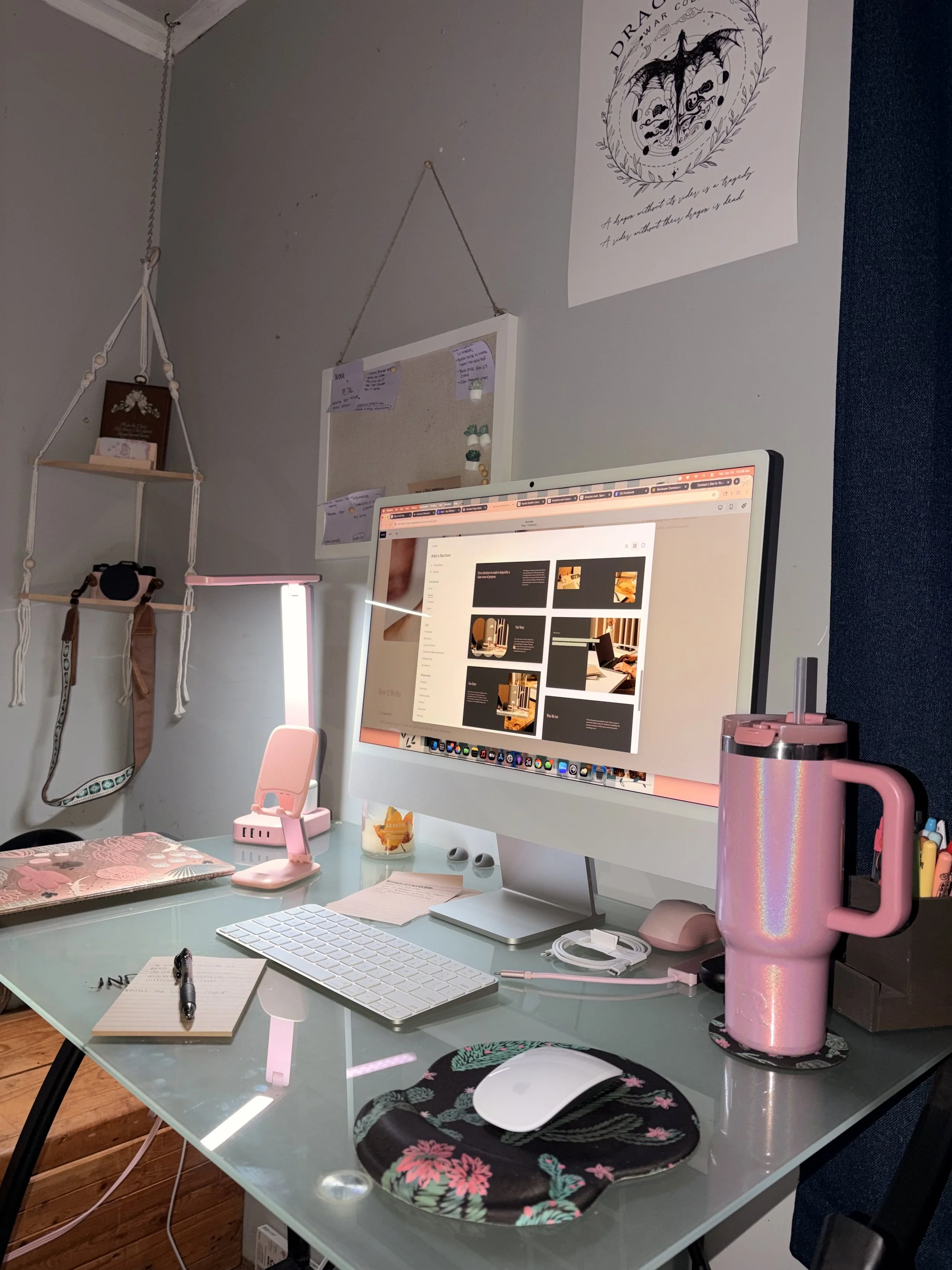 A neatly organized workspace with a glass computer desk holding a desktop computer with a white keyboard, mouse, pink water bottle, notepad, pen, and a pink lamp. Behind the desk, a gray wall features a large Drake poster and a corkboard. To the left, a shelf with a camera and decorative items is hung with white ropes. The workspace is clean with a cozy, organized feel.