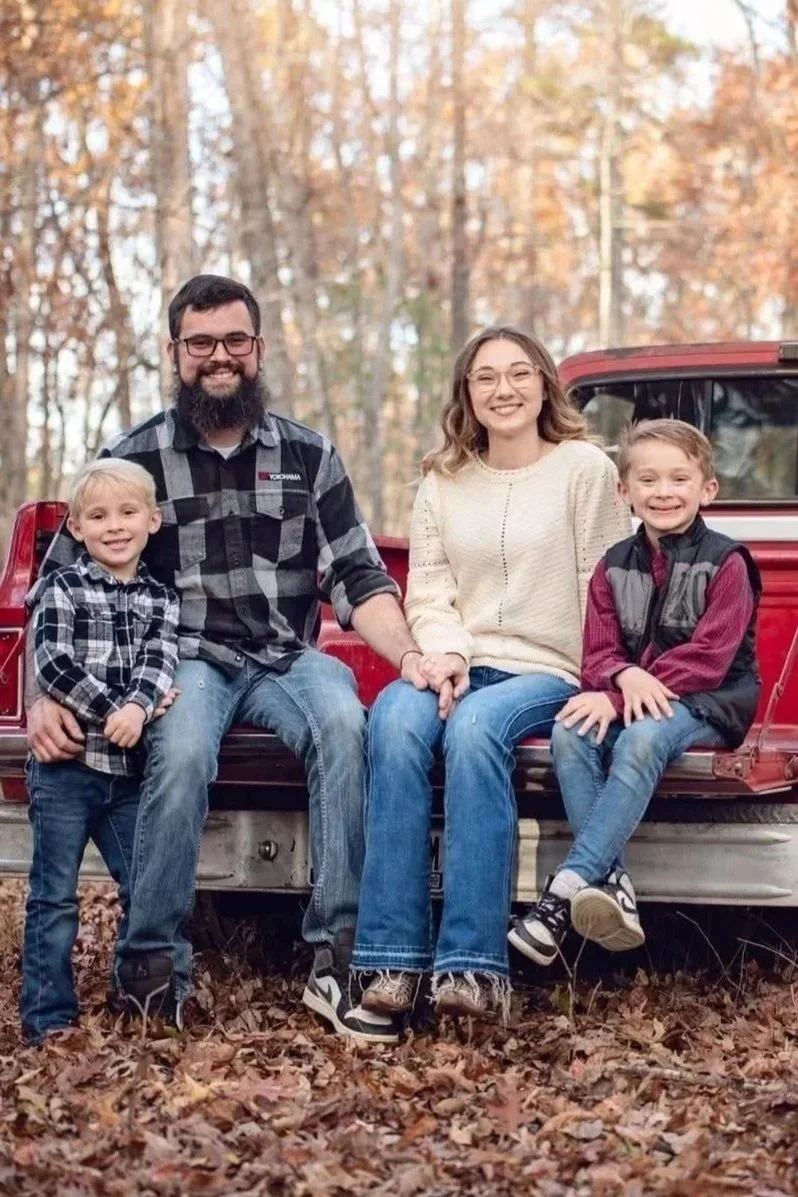 A family of four, including two young boys, a man, and a woman, sitting on the back of a red pickup truck in a wooded area with autumn leaves on the ground and trees with fall foliage.